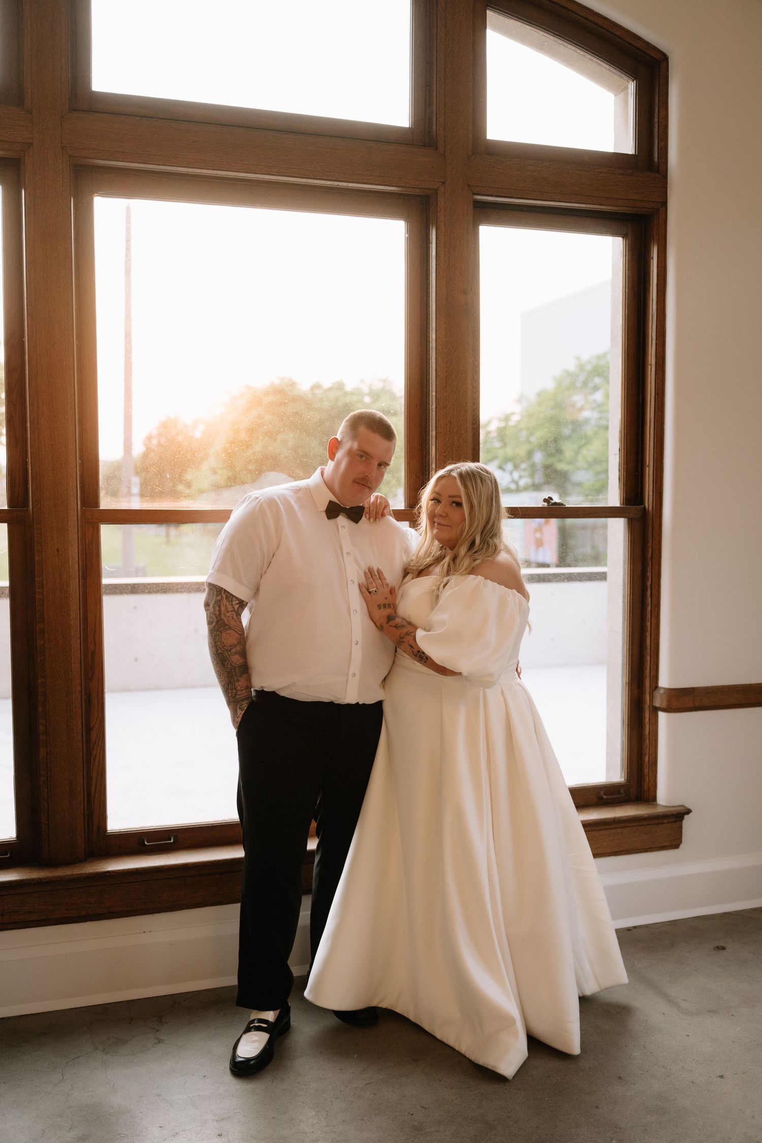 Couple poses before large sunlit windows, groom in bow tie with tattoos, bride in off-shoulder white gown.