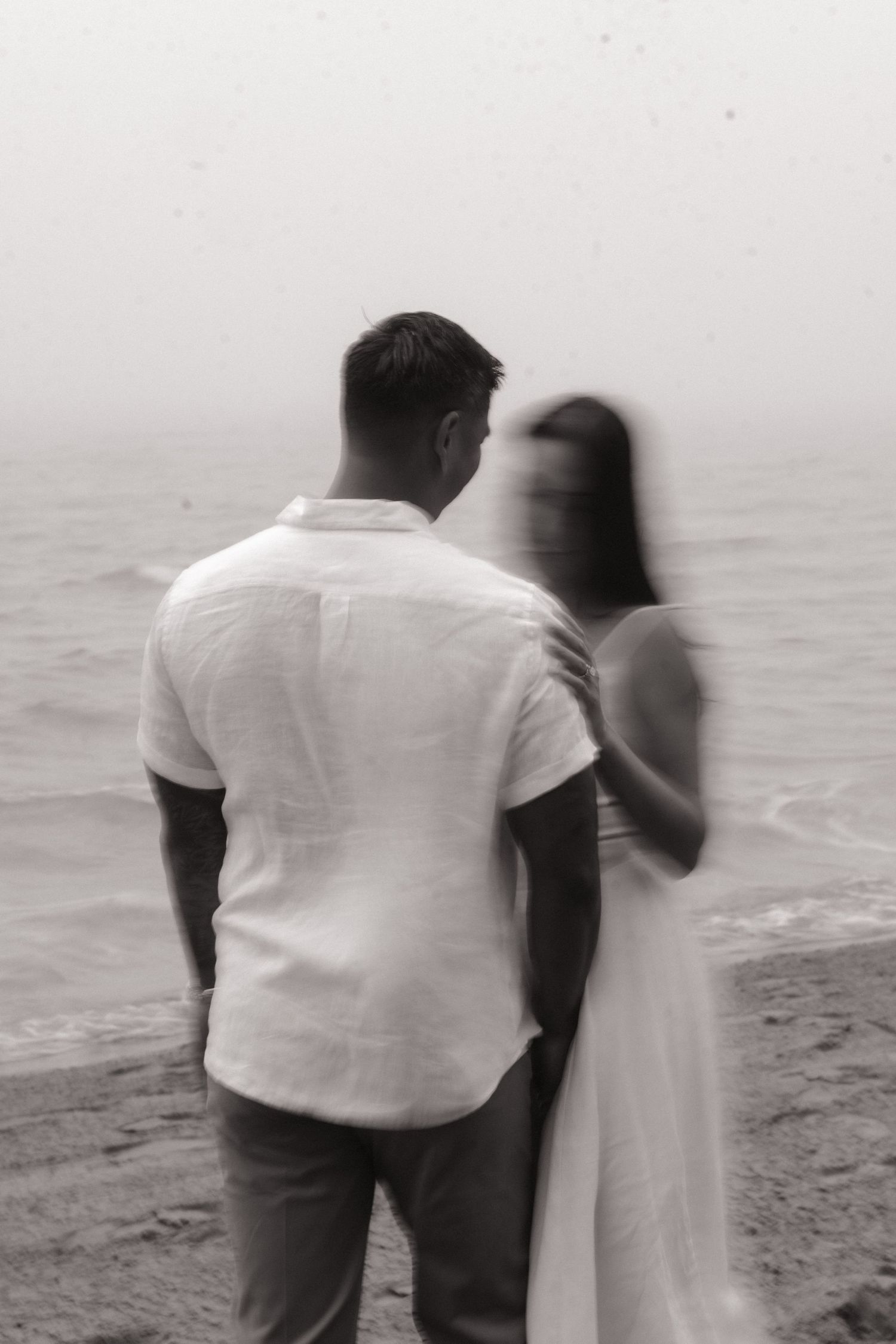 Black and white photo of a couple standing together at the beach, gazing at the ocean waves.