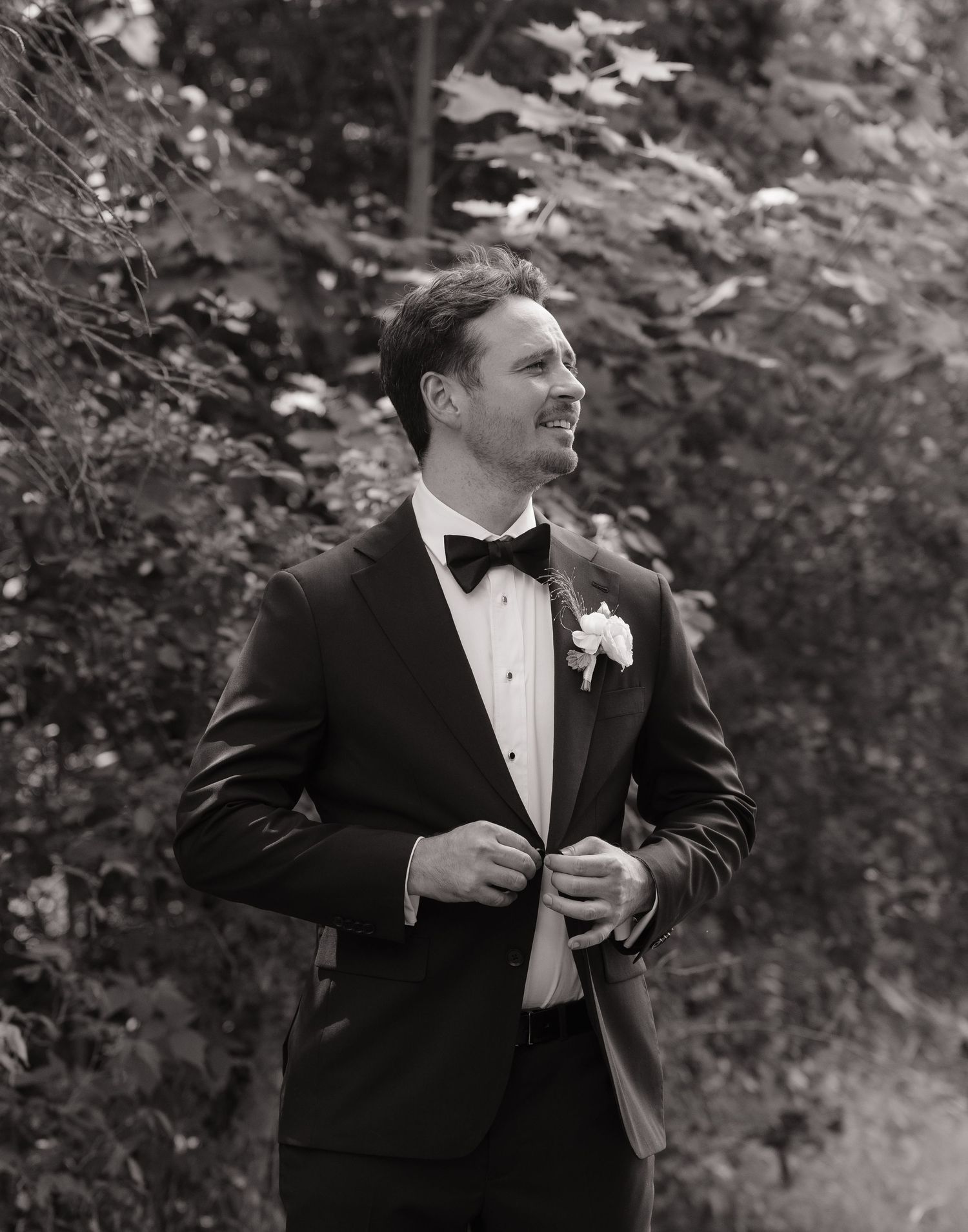 Black and white photo of a groom in a tuxedo with boutonniere, smiling outdoors among lush trees.