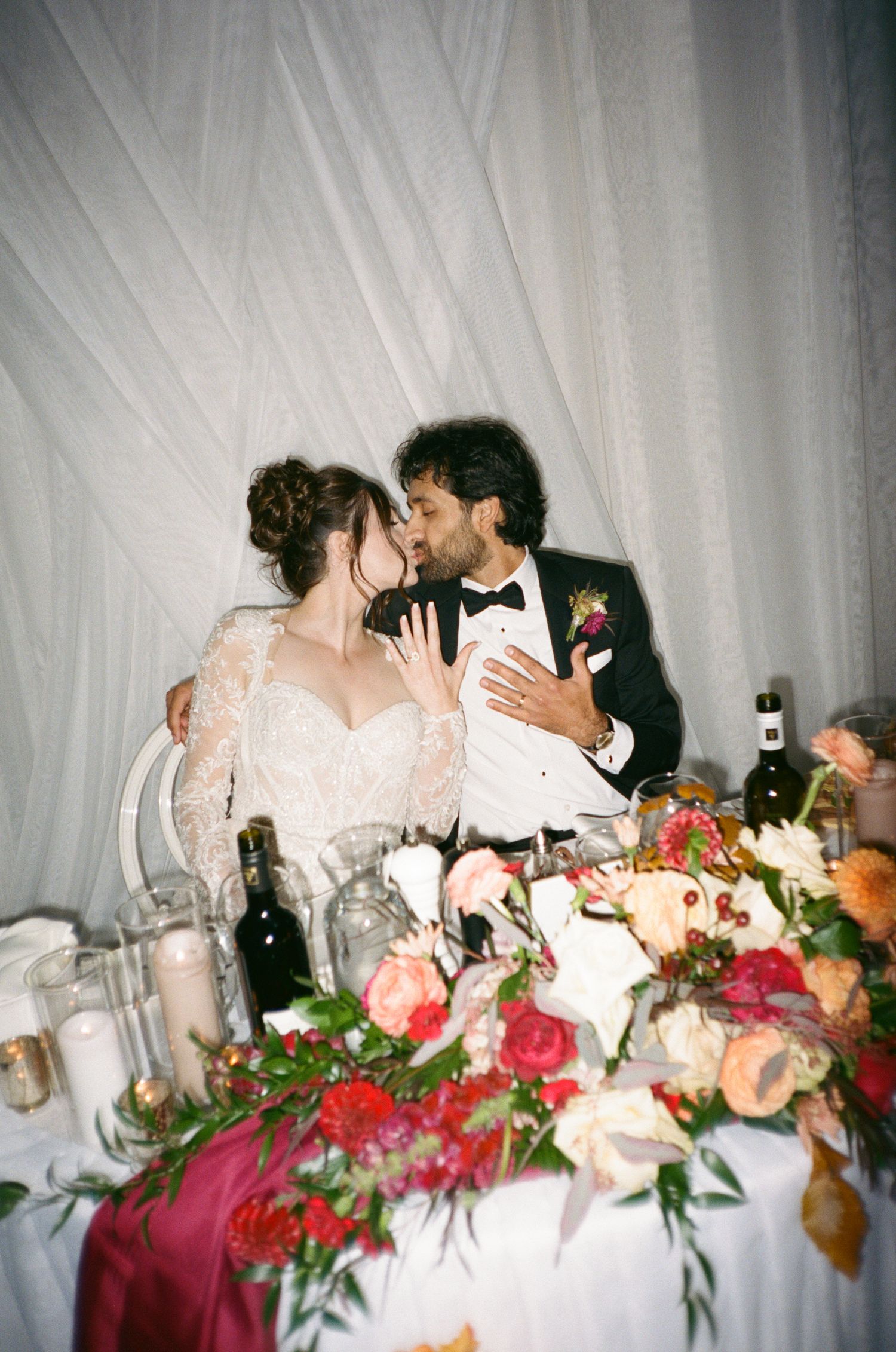 Bride and groom sharing a kiss at sweetheart table adorned with lush floral arrangements at wedding reception.
