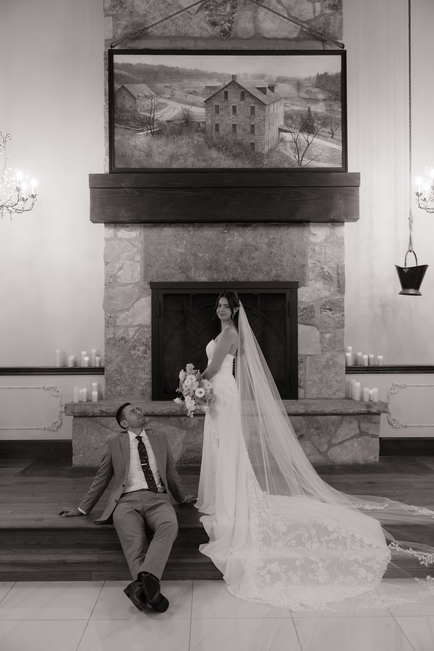 Black and white wedding photo of bride in veil and groom seated by stone fireplace with candles.