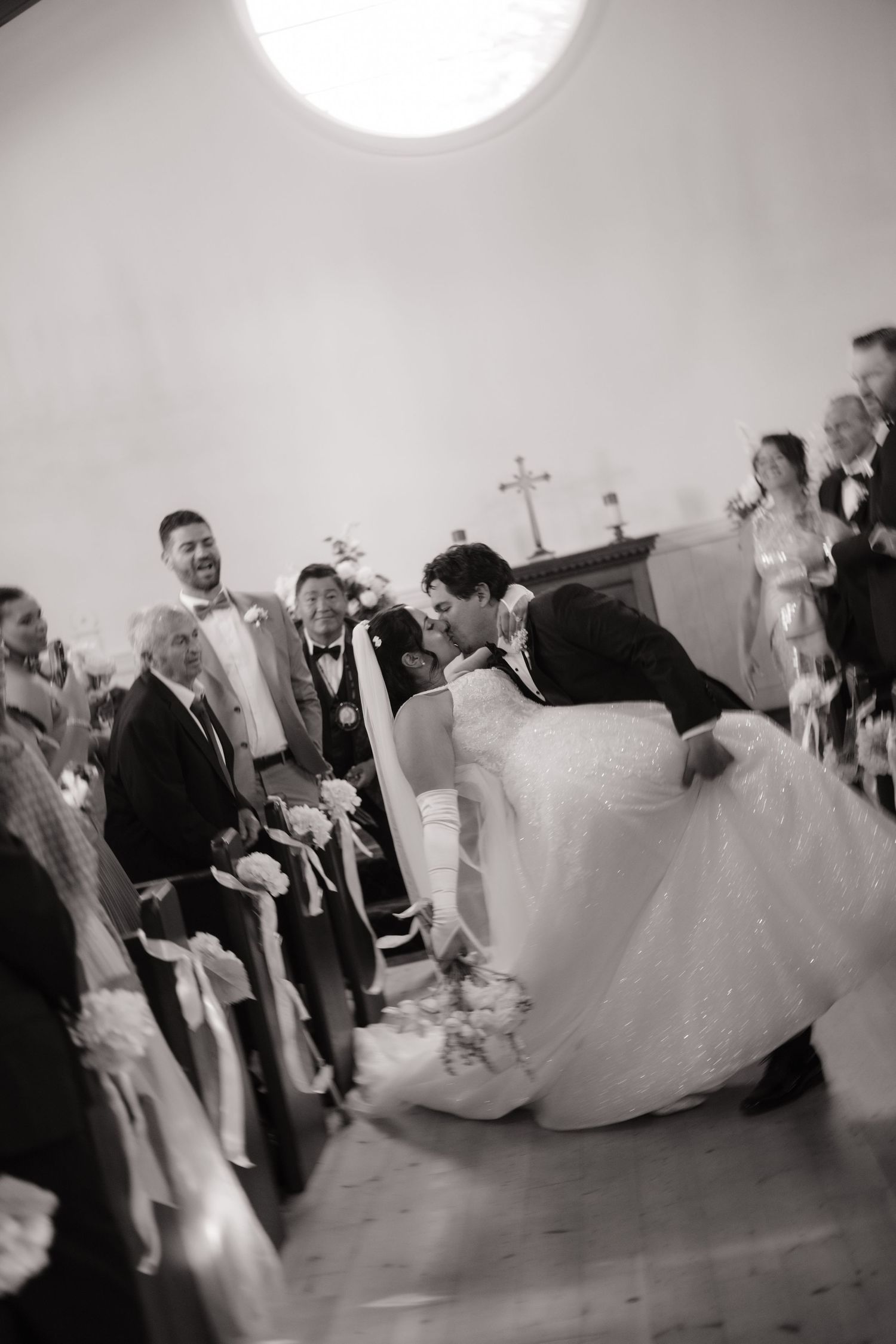 Black and white photo of groom dipping bride for a kiss at the altar as guests cheer.