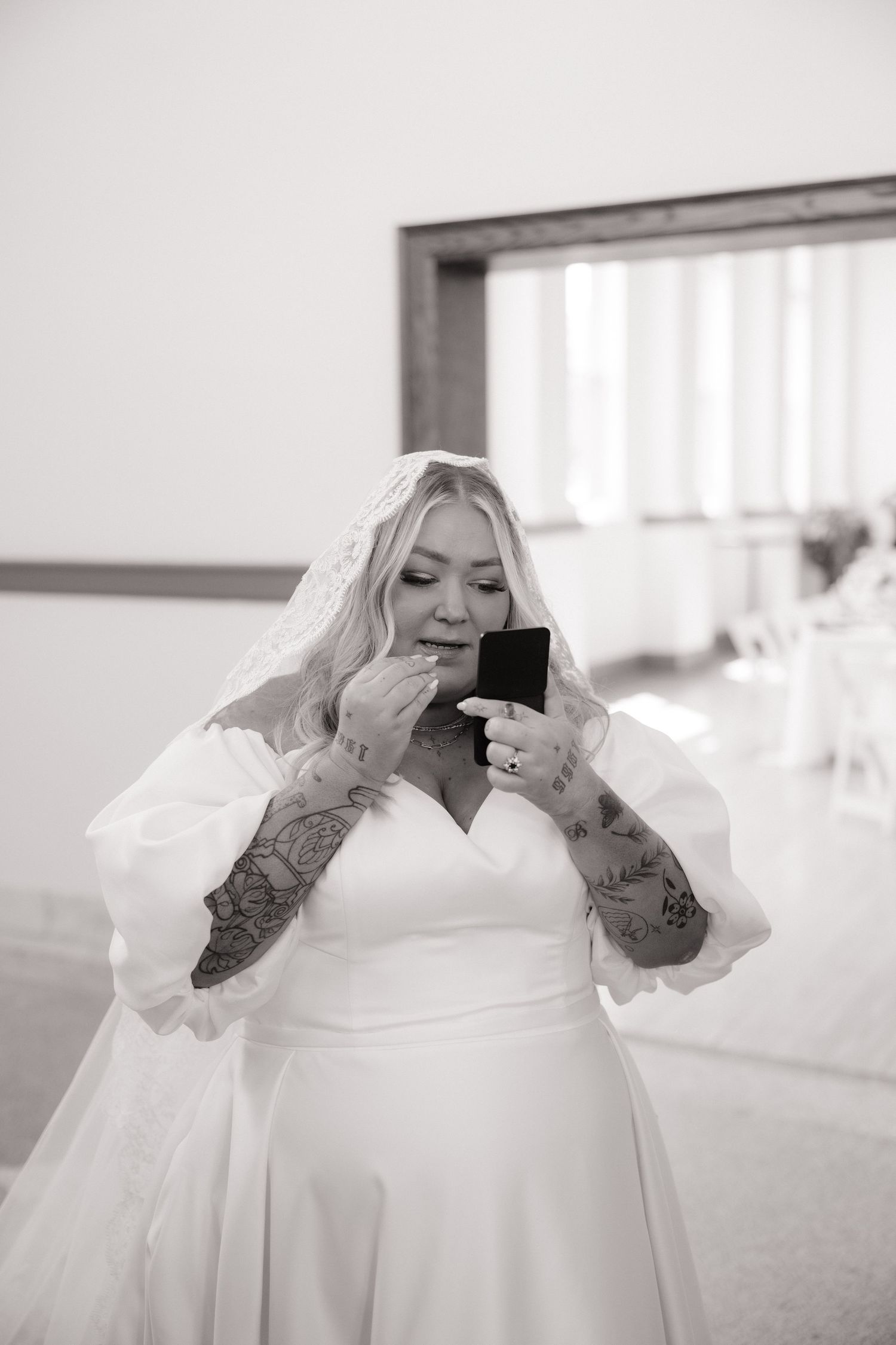 Black and white photo of tattooed bride in white gown and veil touching up makeup with compact mirror.