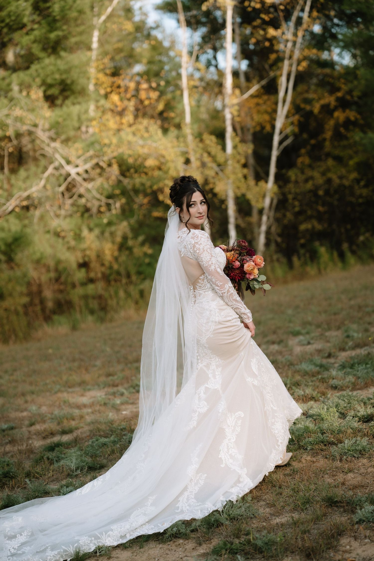 Bride in lace mermaid gown with cathedral veil holds autumn bouquet in golden forest setting.