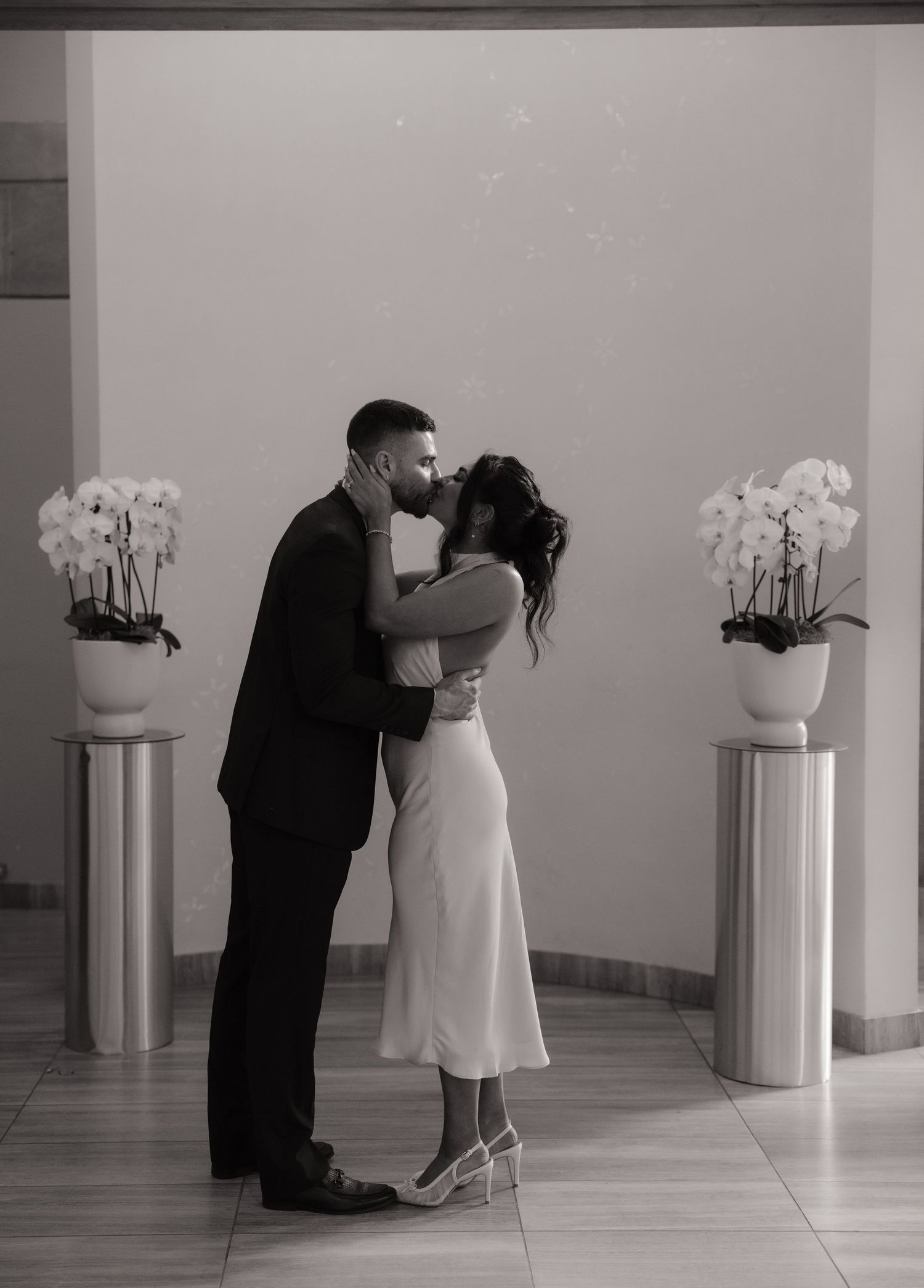 Black and white photo of newlyweds sharing a kiss beside white orchid arrangements indoors.