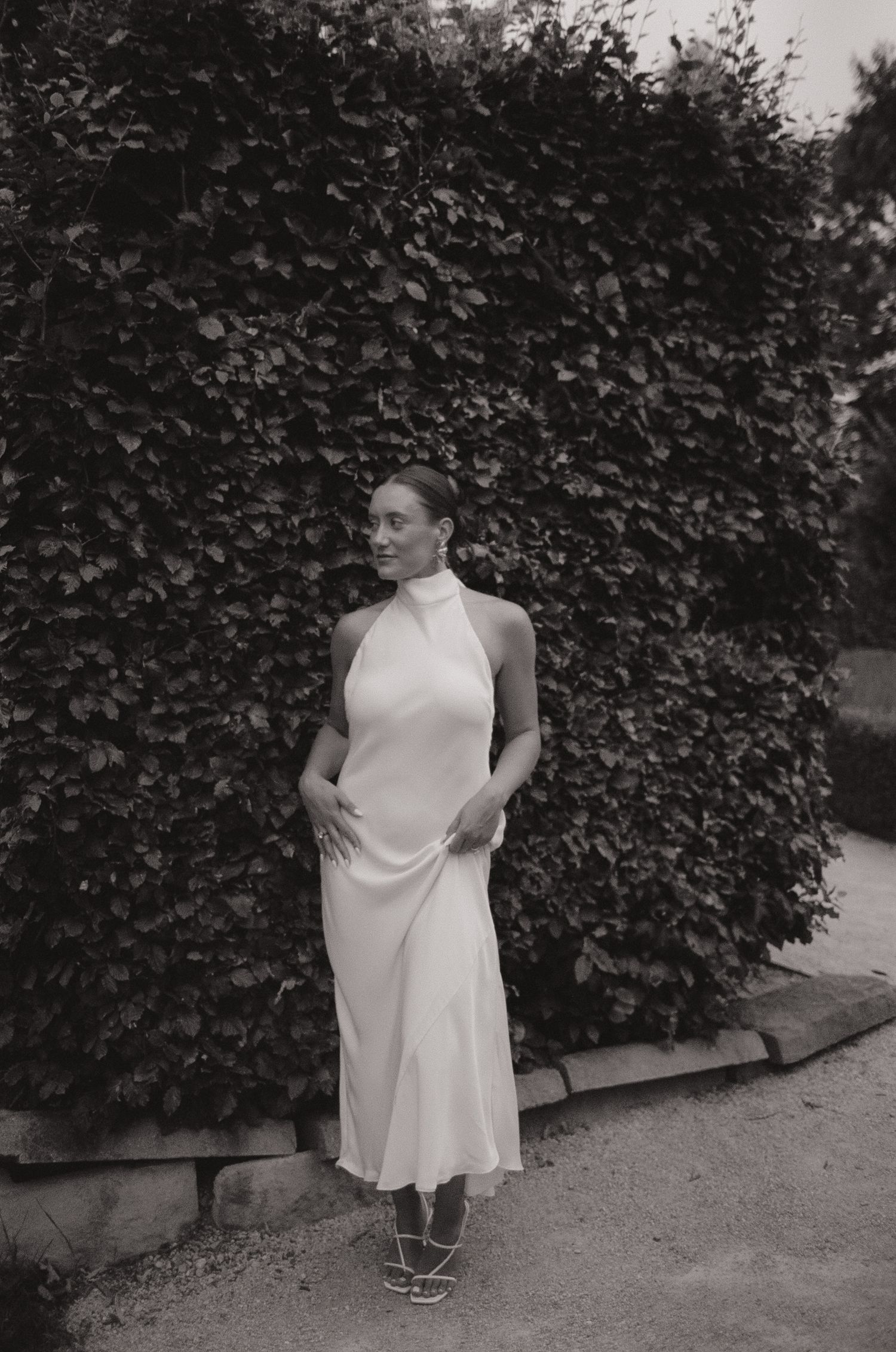 Woman in white halter midi dress and strappy sandals posing before lush green hedge in black and white photo.