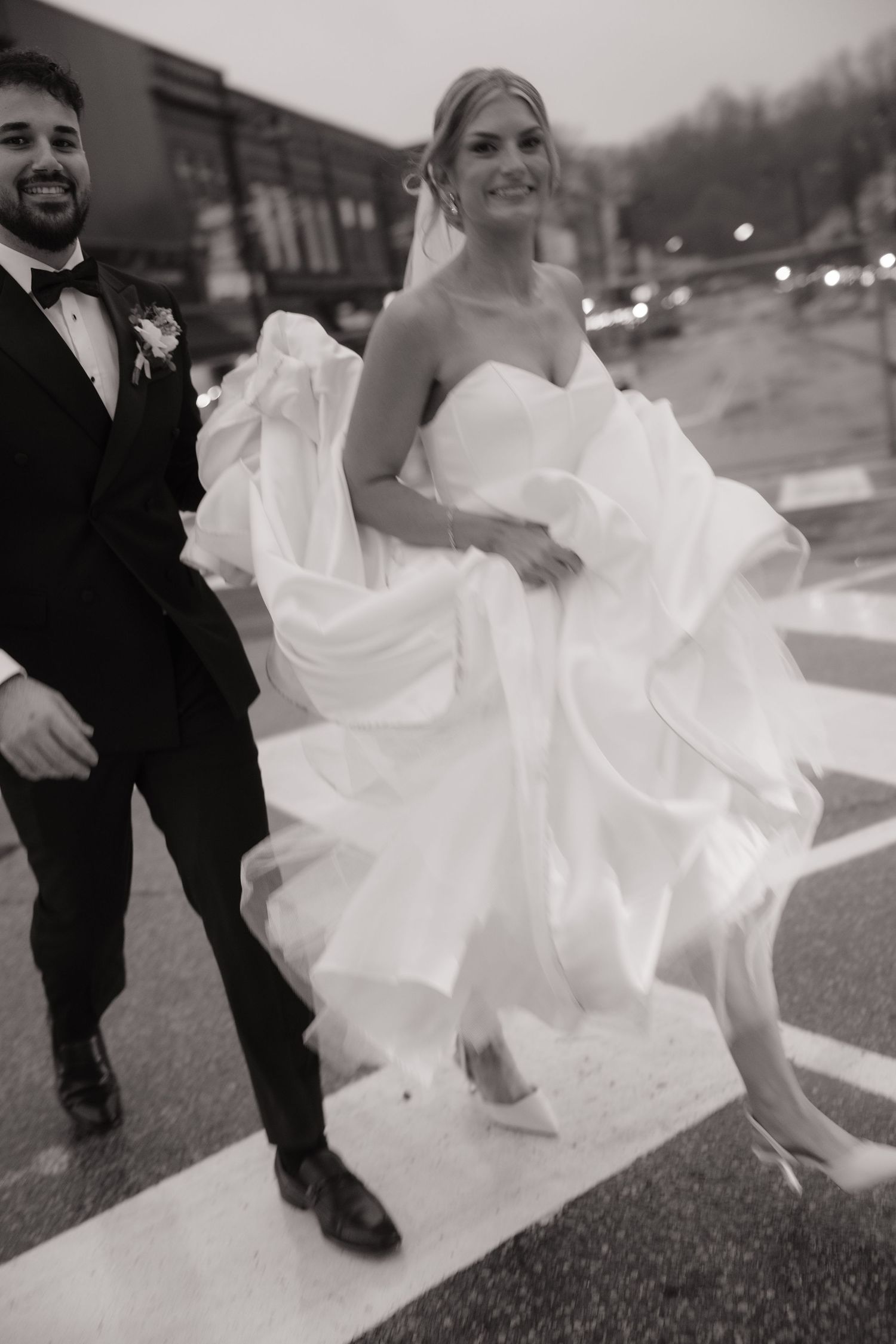 Black and white candid of smiling bride lifting flowing white gown while crossing street with groom.