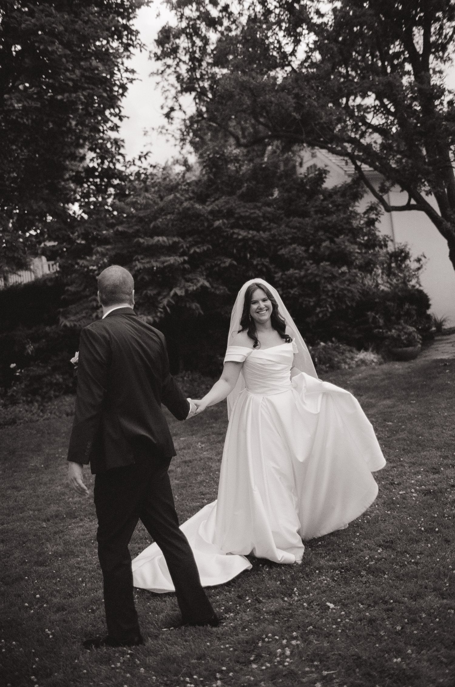 Black and white photo of a bride in a white gown and veil holding hands with a groom in a suit outdoors.