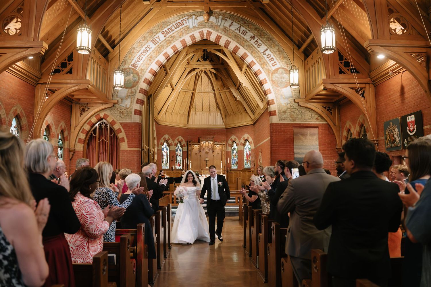 Bride and groom walk down the aisle of a stunning wooden chapel surrounded by applauding wedding guests.