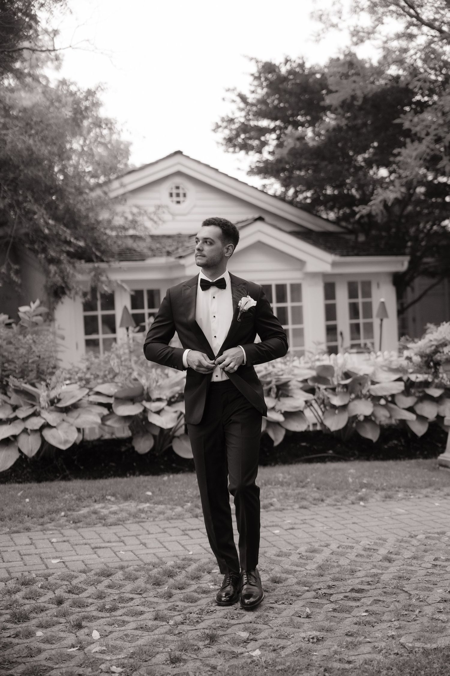 Groom in black tuxedo with bow tie buttoning jacket outside elegant white house surrounded by lush greenery.