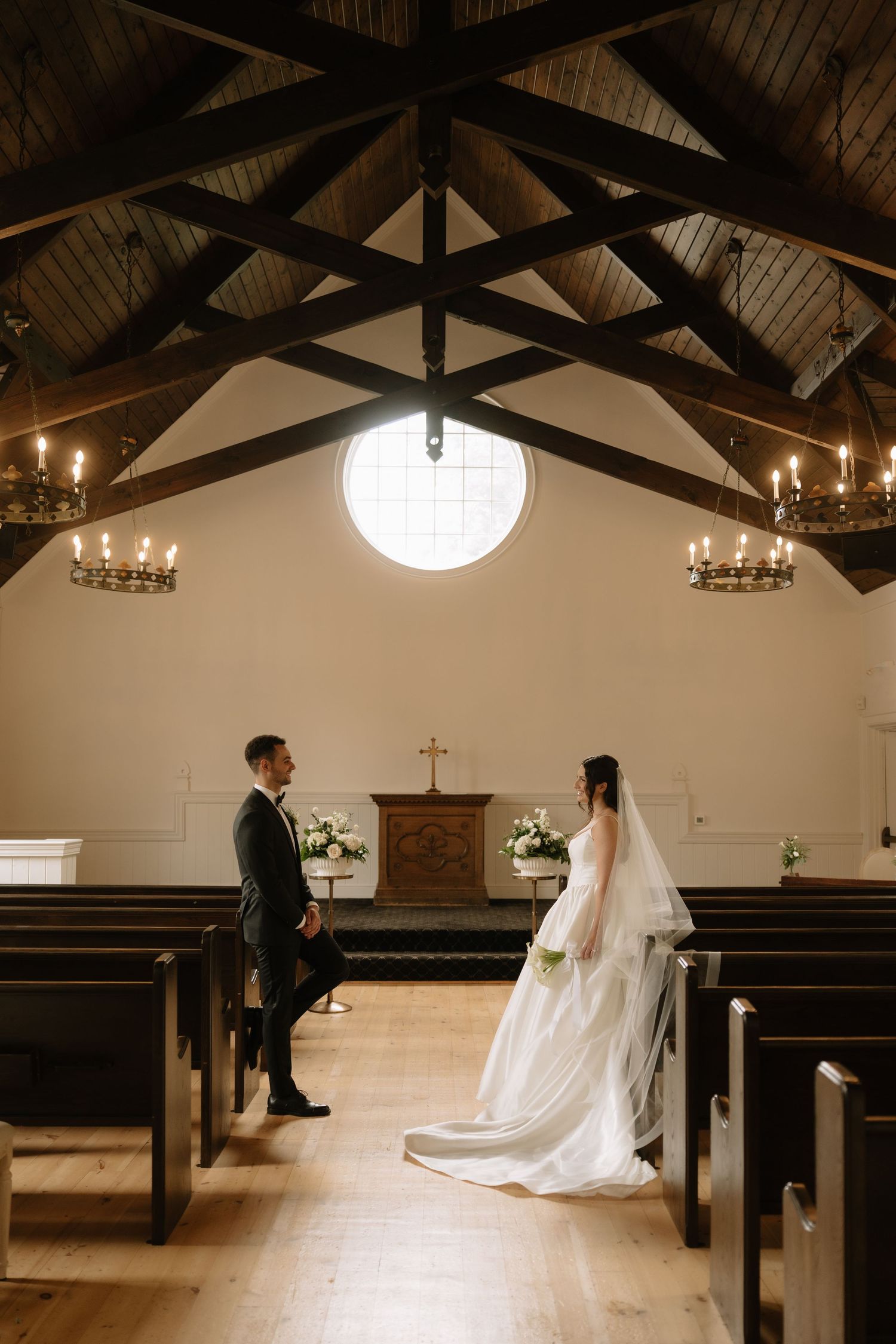 Bride and groom facing each other in a charming white chapel with wooden beam ceiling and candlelit chandeliers.