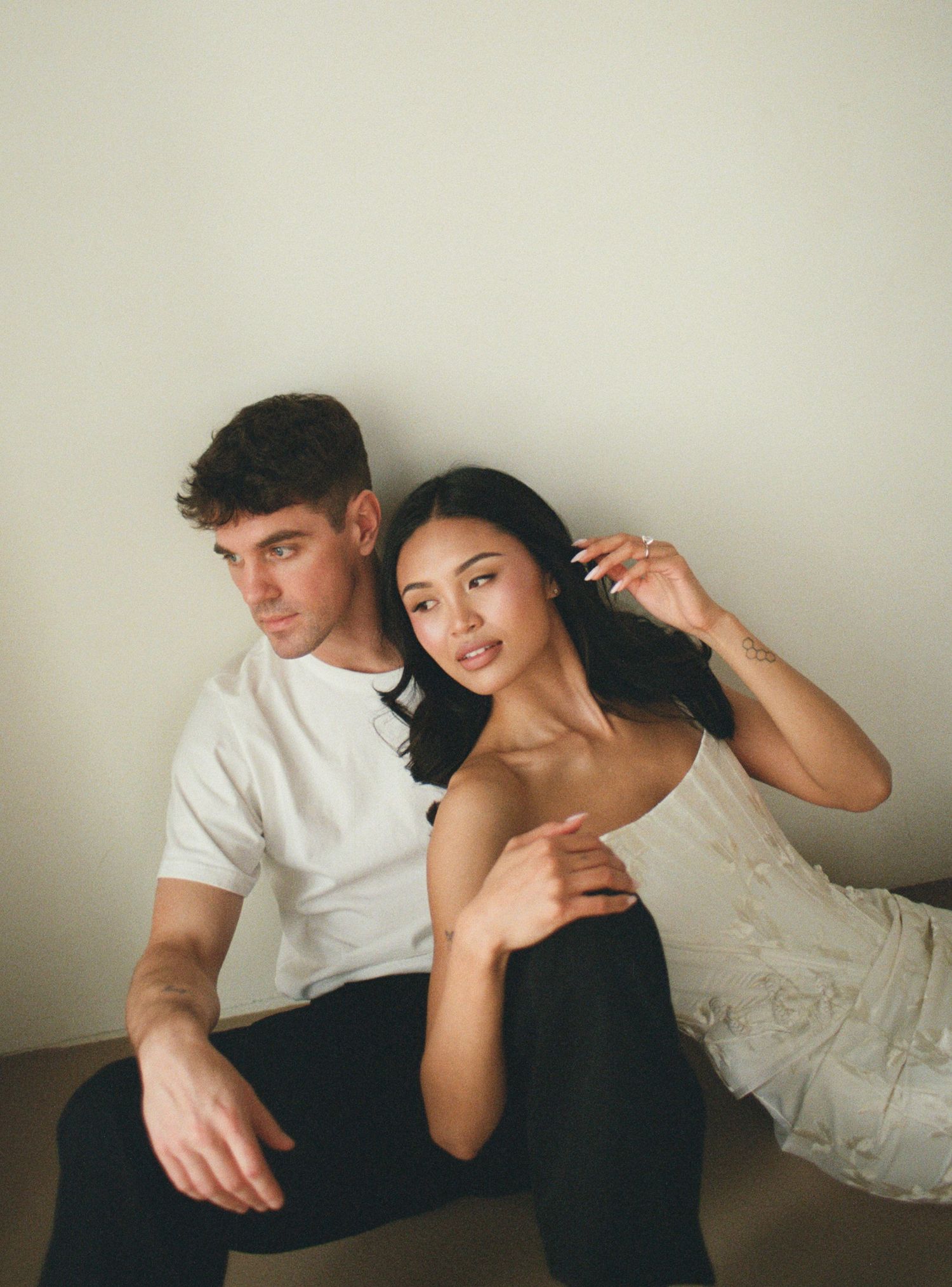 Stylish couple sitting against a white wall, woman in lace dress leaning on mans shoulder.