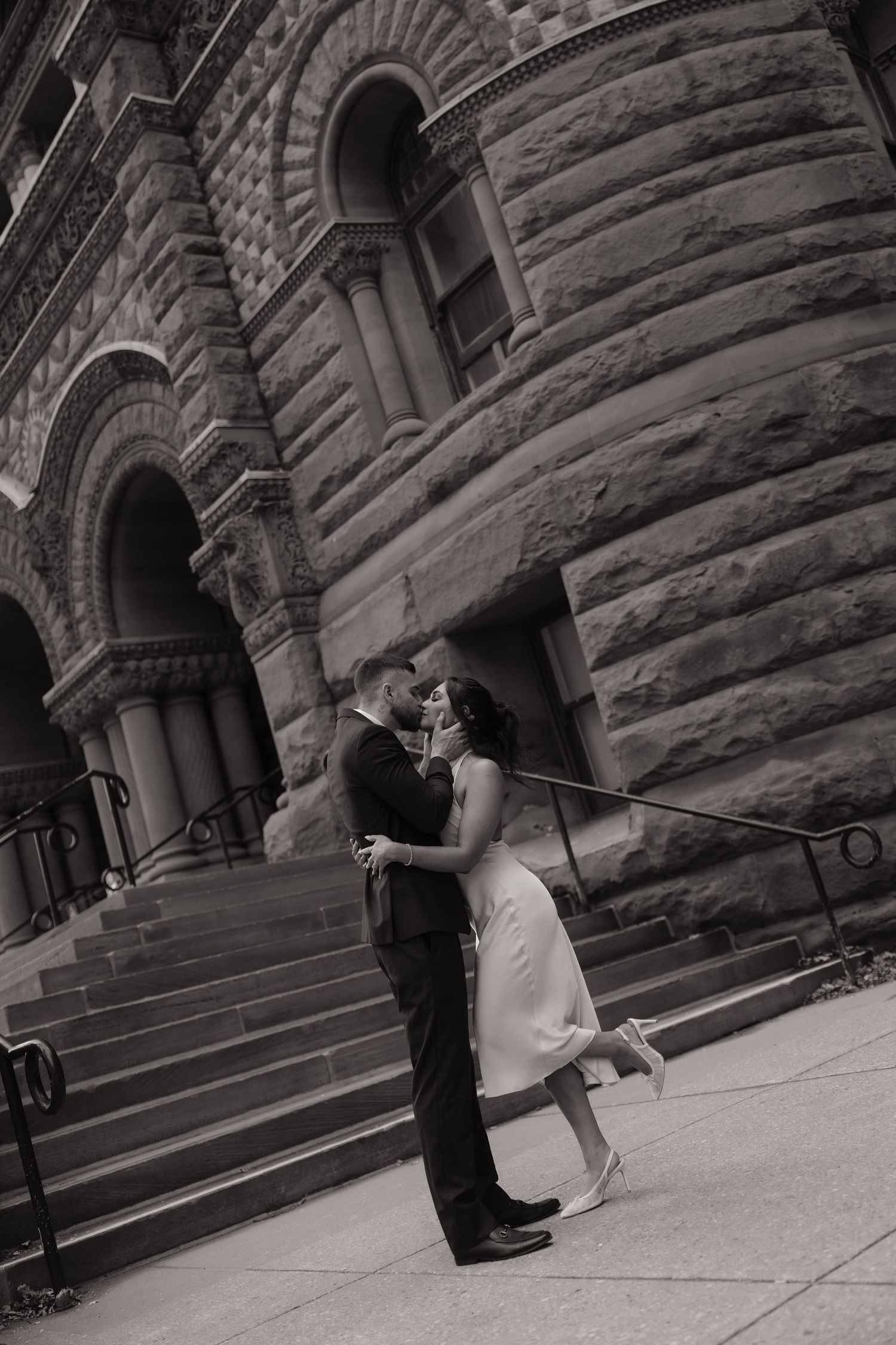 Black and white romantic kiss between bride and groom on steps of a grand historic stone building.