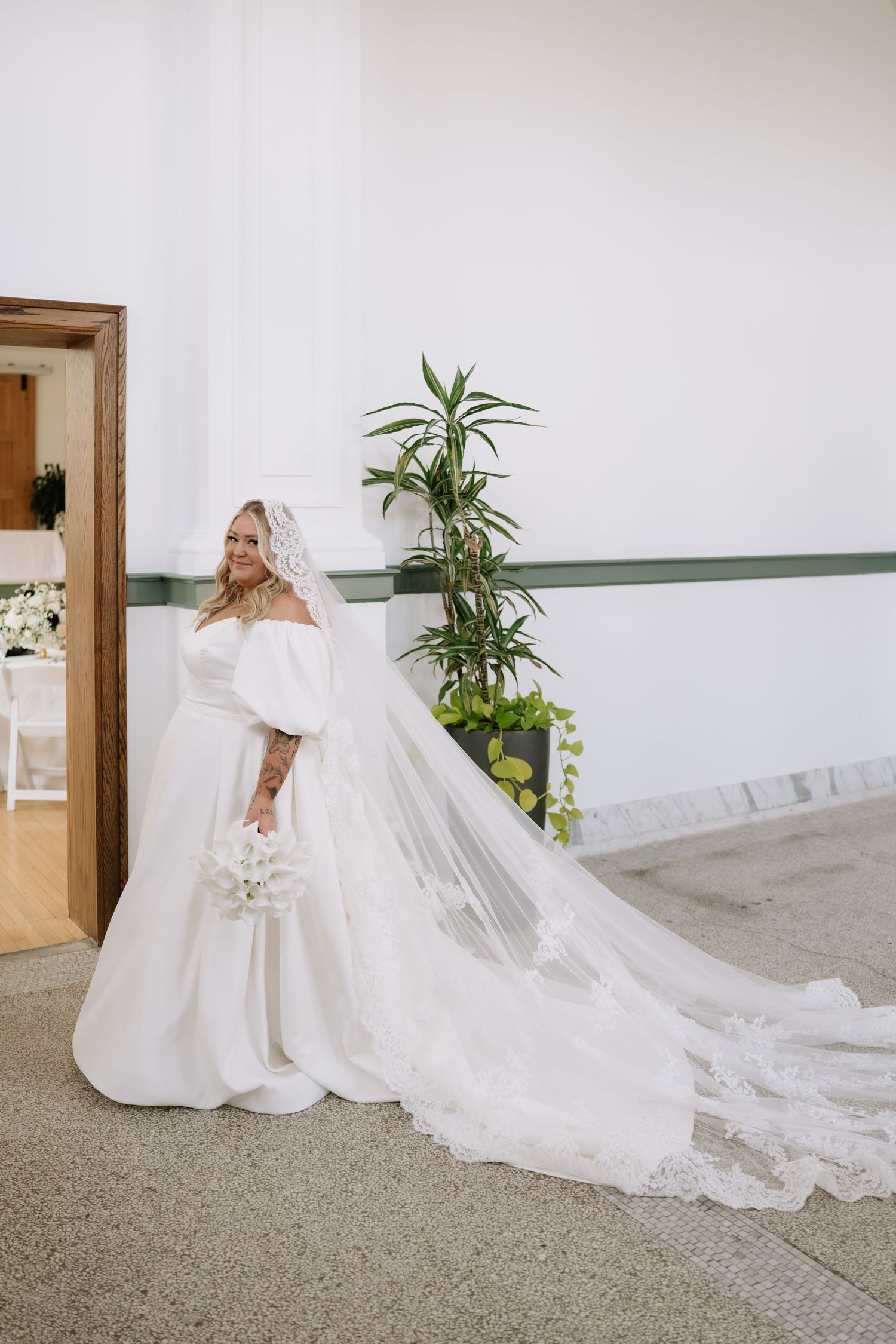 Bride in off-shoulder white ball gown with cathedral veil and bouquet standing in elegant venue hallway.