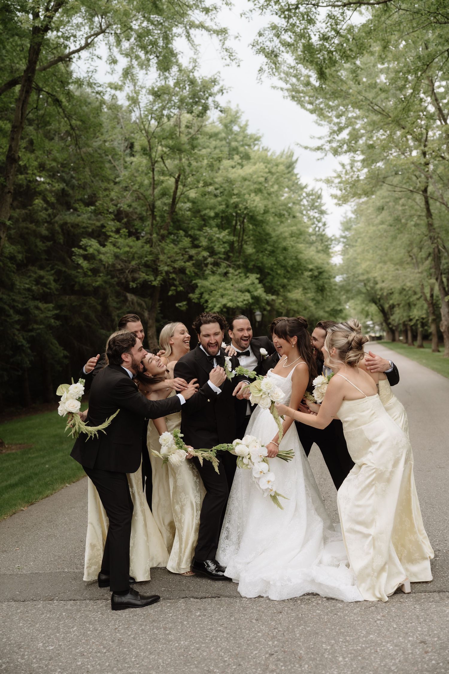 Wedding party in black tuxedos and champagne gowns laughing together on a tree-lined path with the bride and groom.