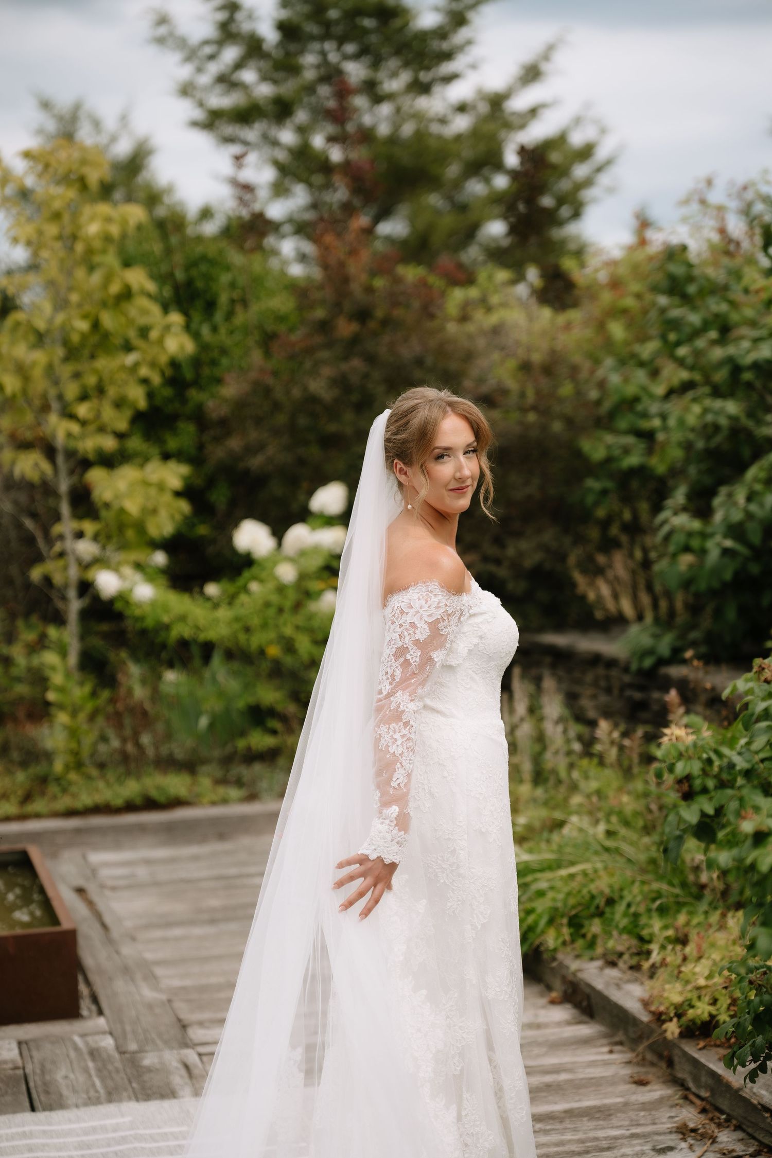 Bride in off-shoulder lace gown with cathedral veil poses gracefully on outdoor wooden deck surrounded by greenery.