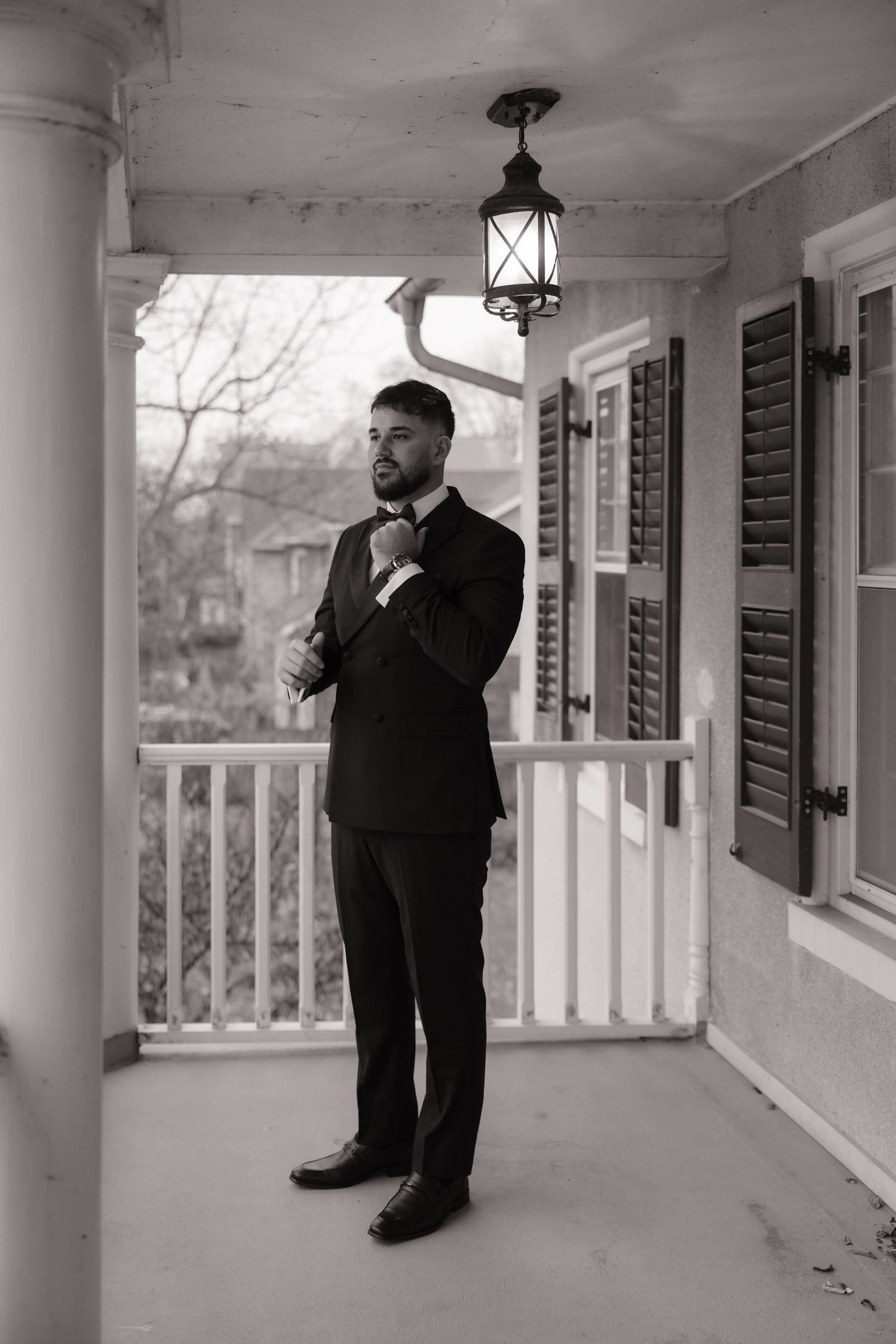 Black and white portrait of dapper groom in tuxedo adjusting bow tie on a classic home porch.