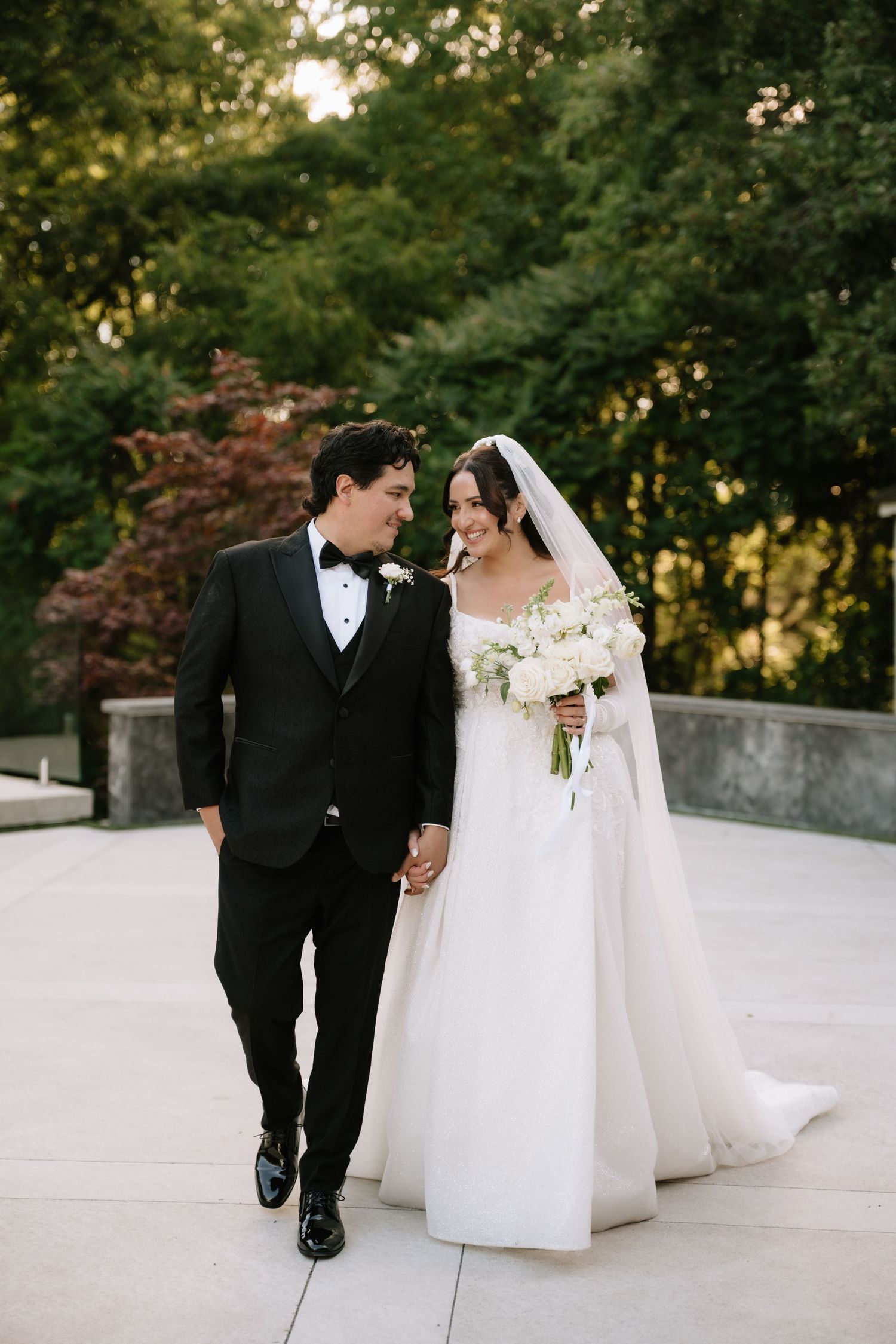 Newlyweds walk hand in hand on stone terrace, bride carries white bouquet in elegant gown.