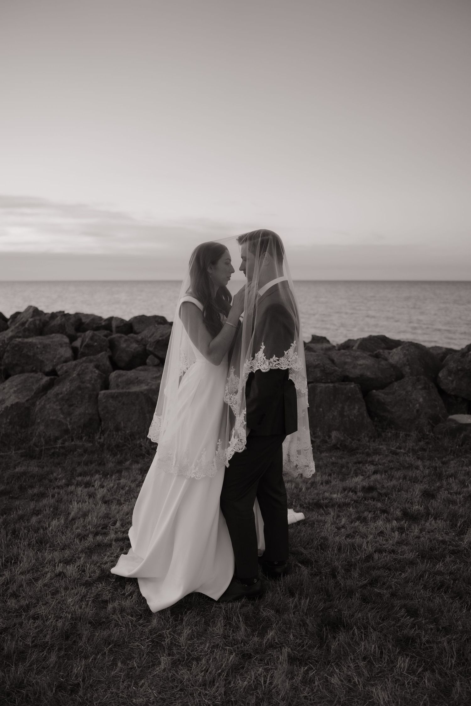 Bride and groom share intimate moment by ocean rocks at dusk in black and white.