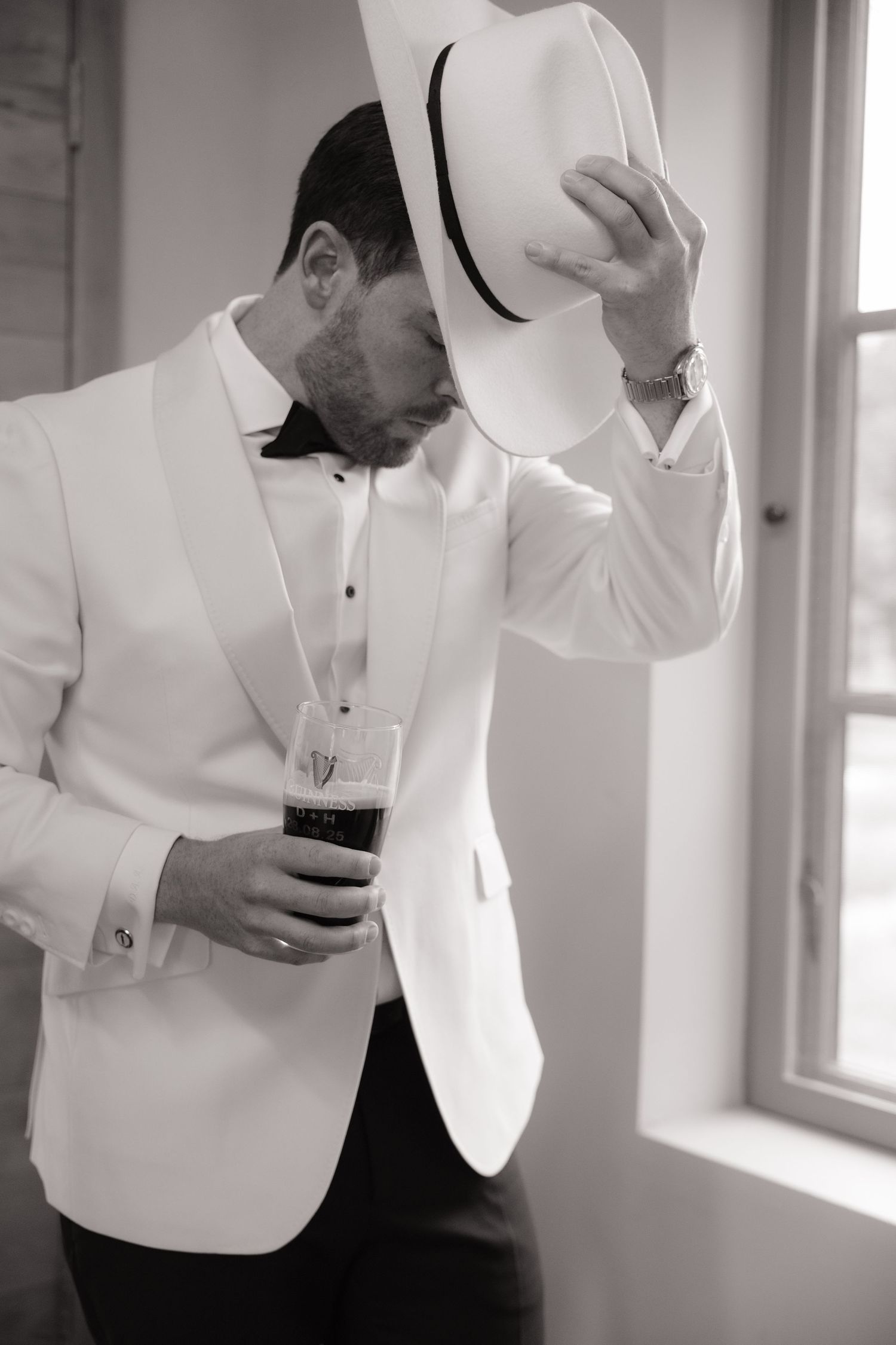 Groom in white tuxedo jacket and fedora hat holding a dark beer glass indoors.