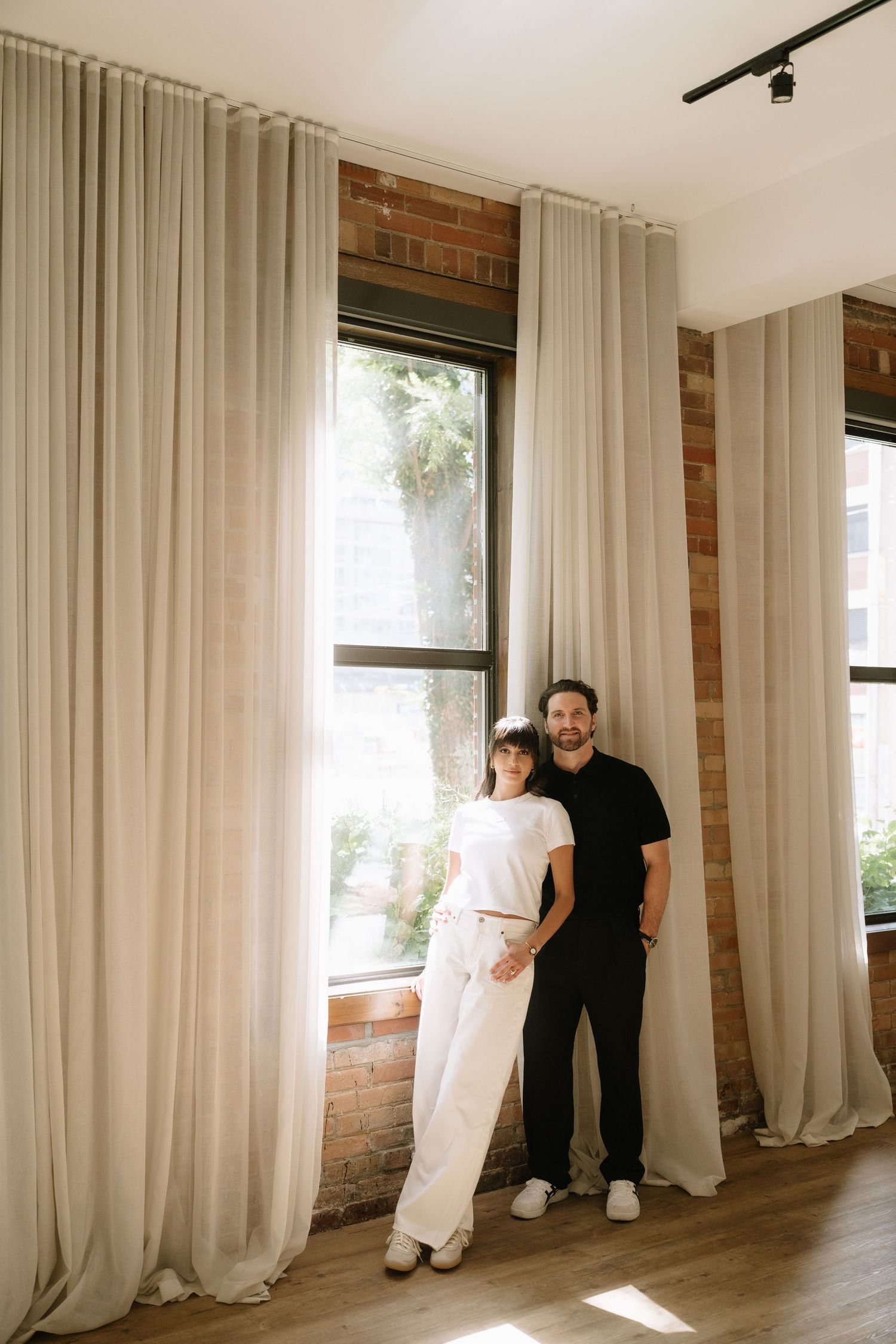 Couple posing together by large windows with flowing linen curtains in a modern brick-accented space.