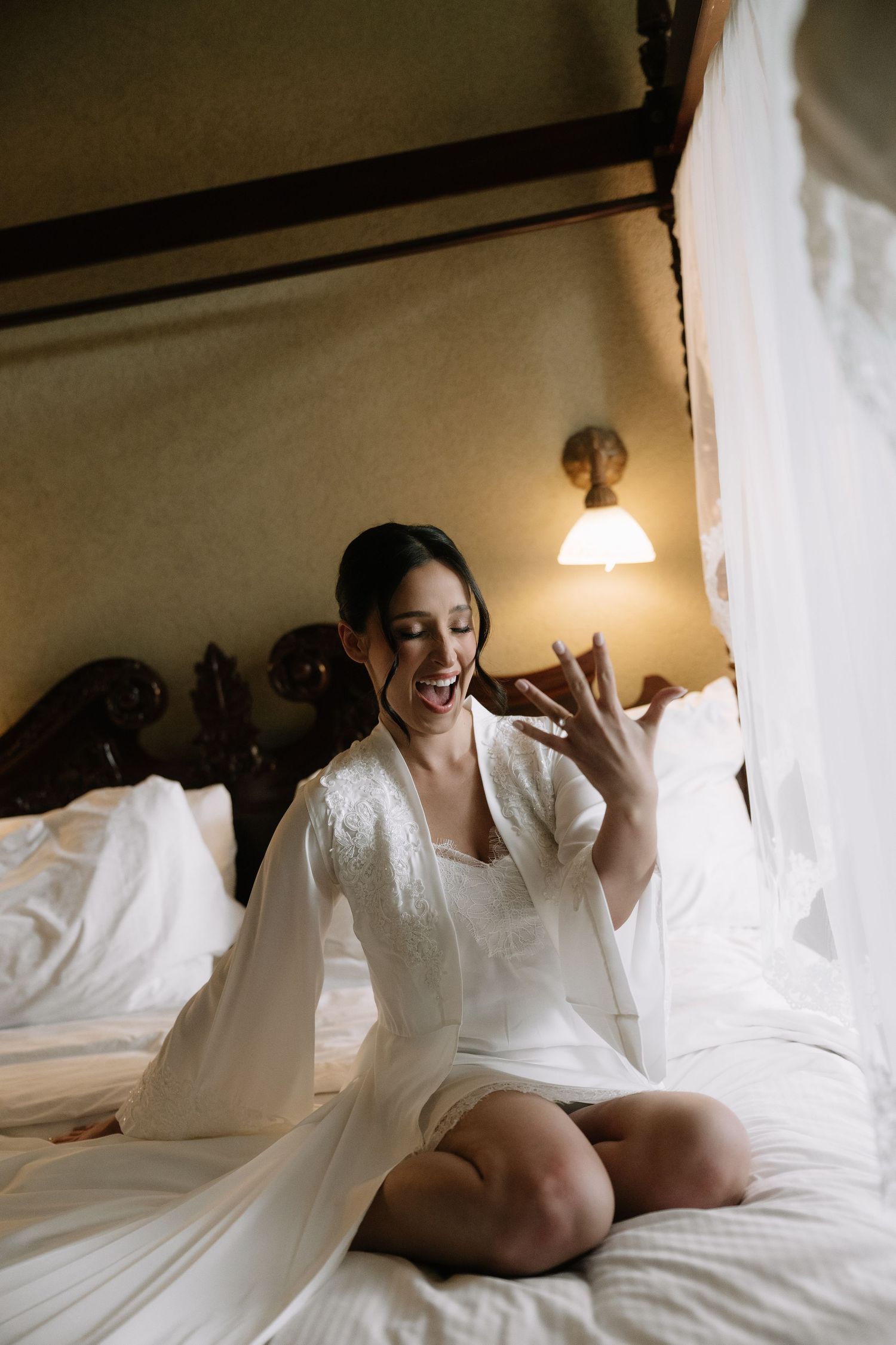 Excited bride in white lace robe sitting on bed admiring her engagement ring.