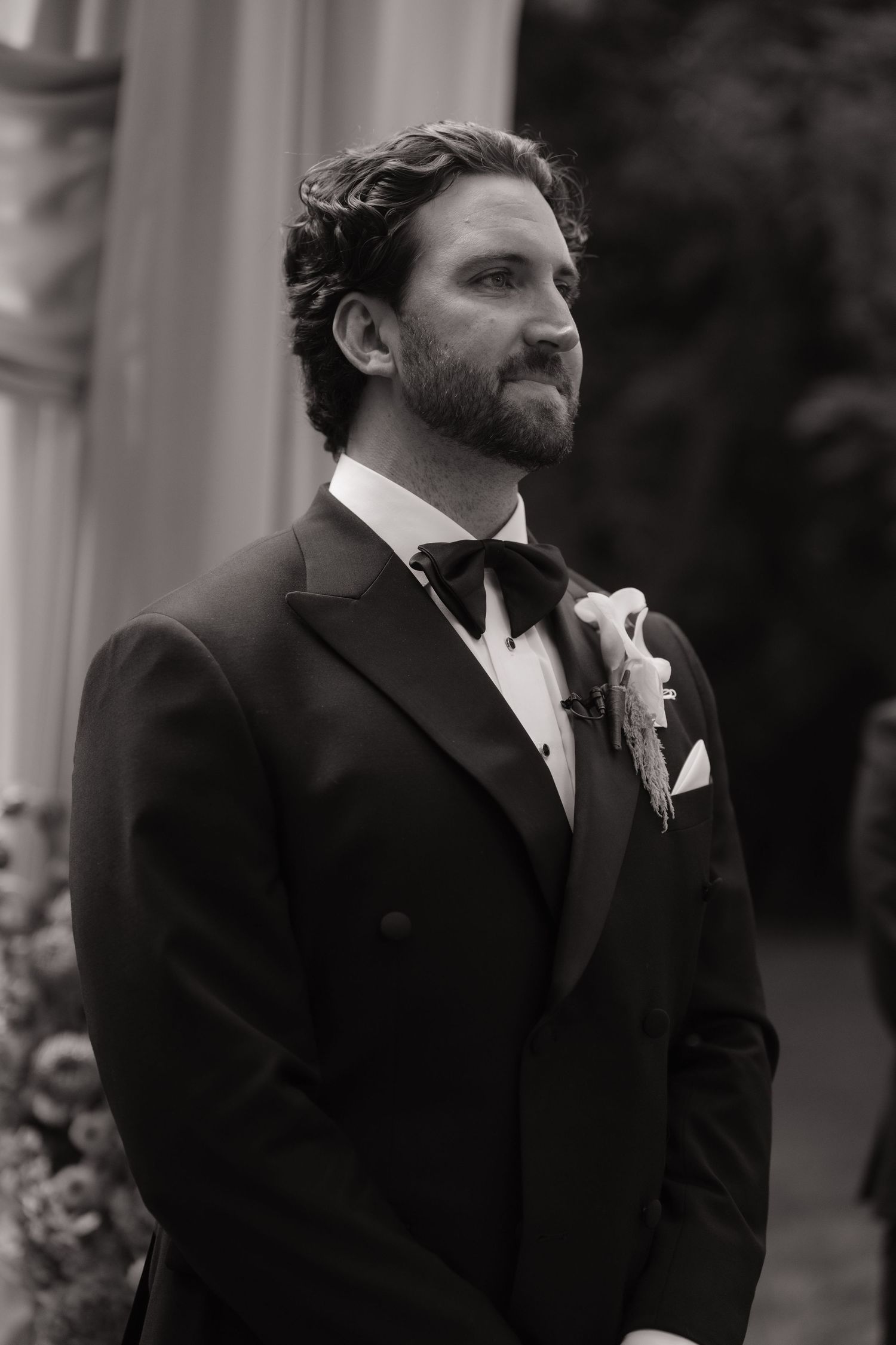 Black and white photo of a groom in a tuxedo with boutonniere, gazing forward at his wedding ceremony.