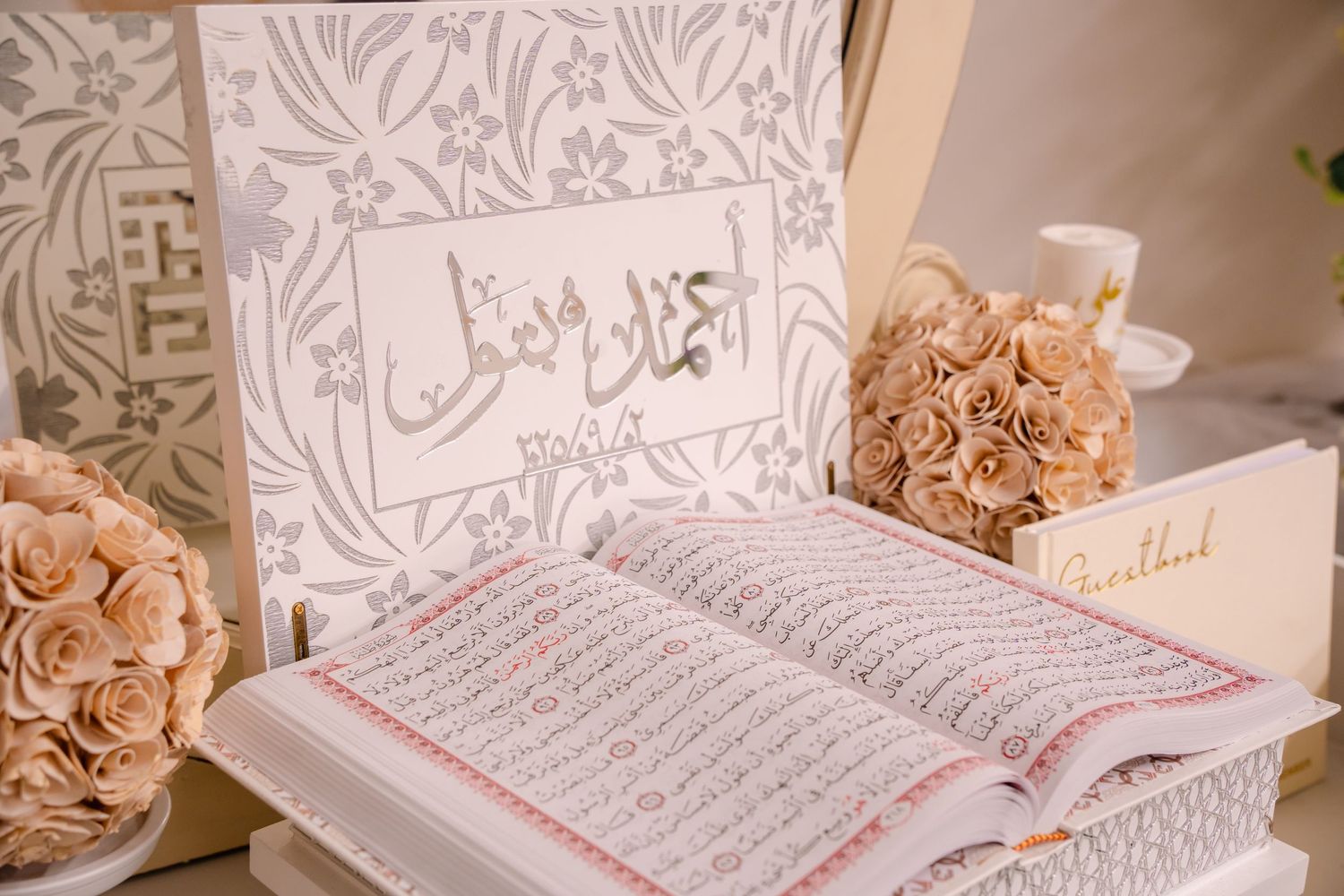 open Quran, Ahmad and Batool written in arabic on a white sign with metallic silver text, small beige bouquet of flower on either side.