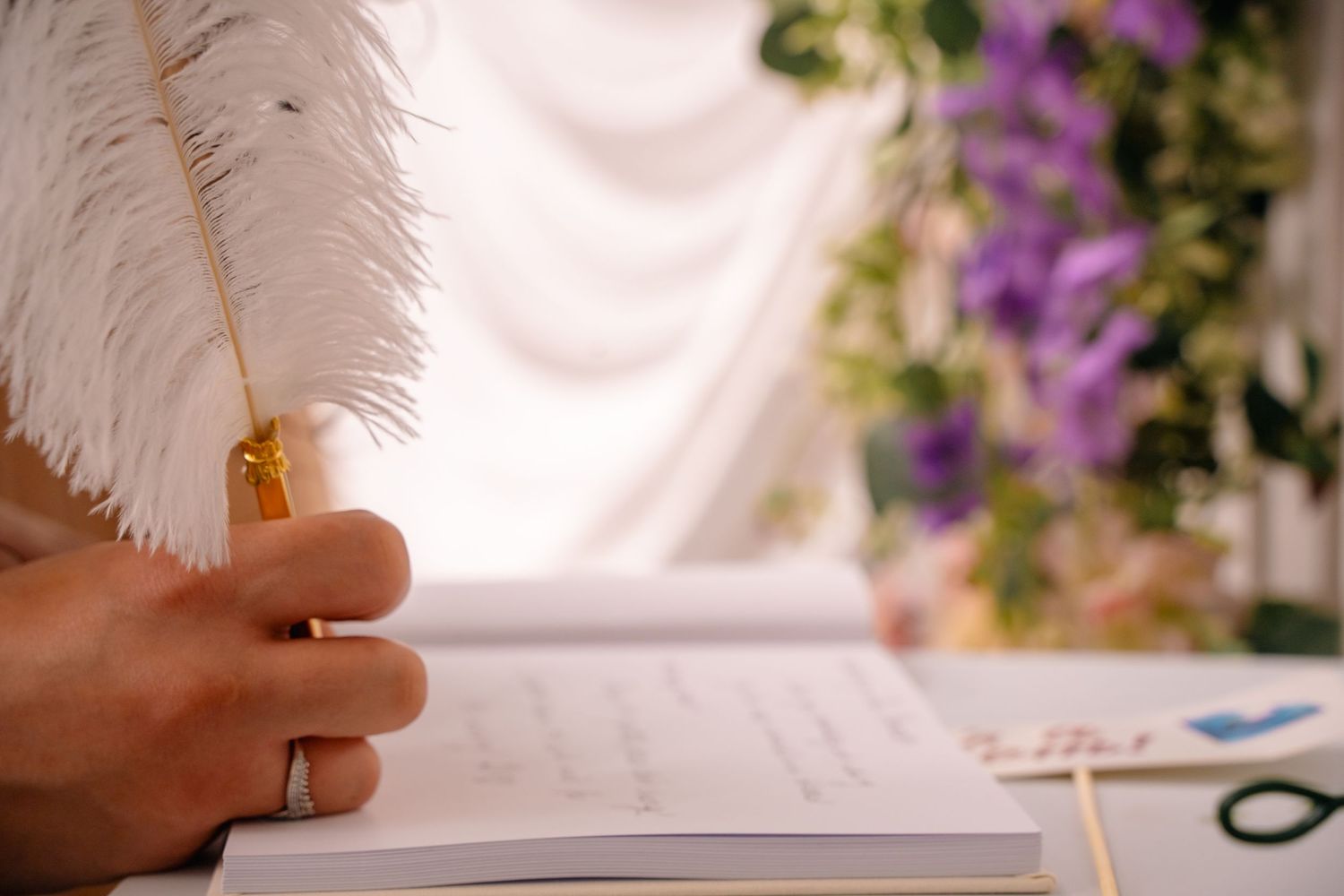 hand writing with a feather pen in guestbook, with purple flowers and green leaves in the background. Tied in Time Photography.