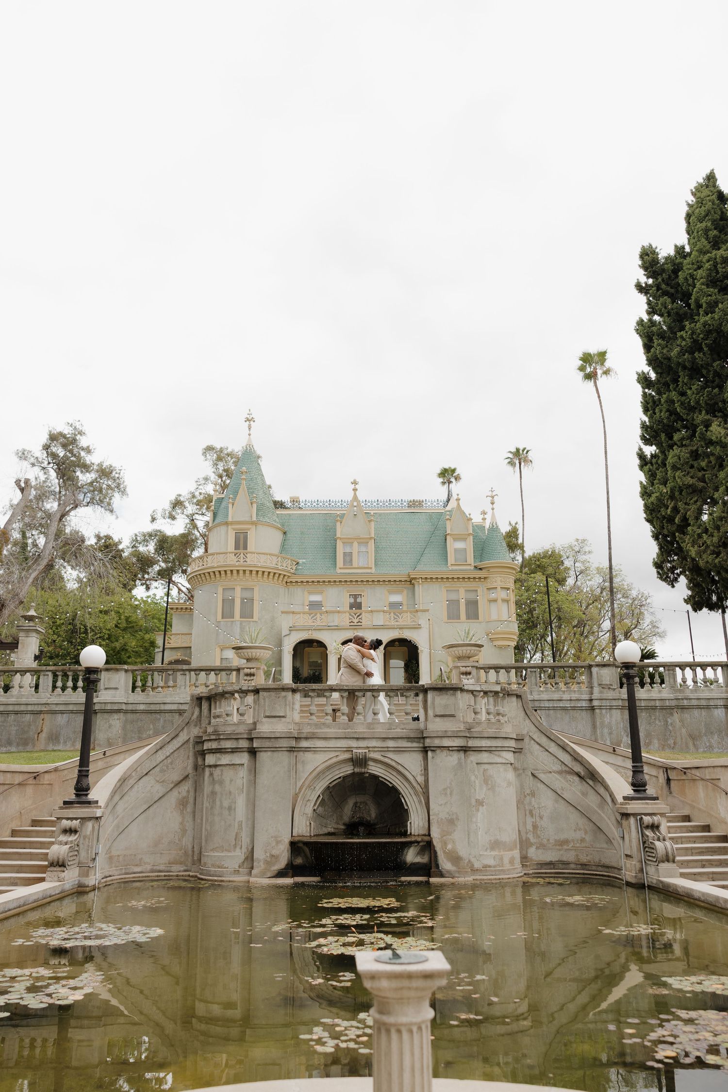 Couple embracing on grand staircase of elegant castle venue with lily pond reflecting ornate architecture.