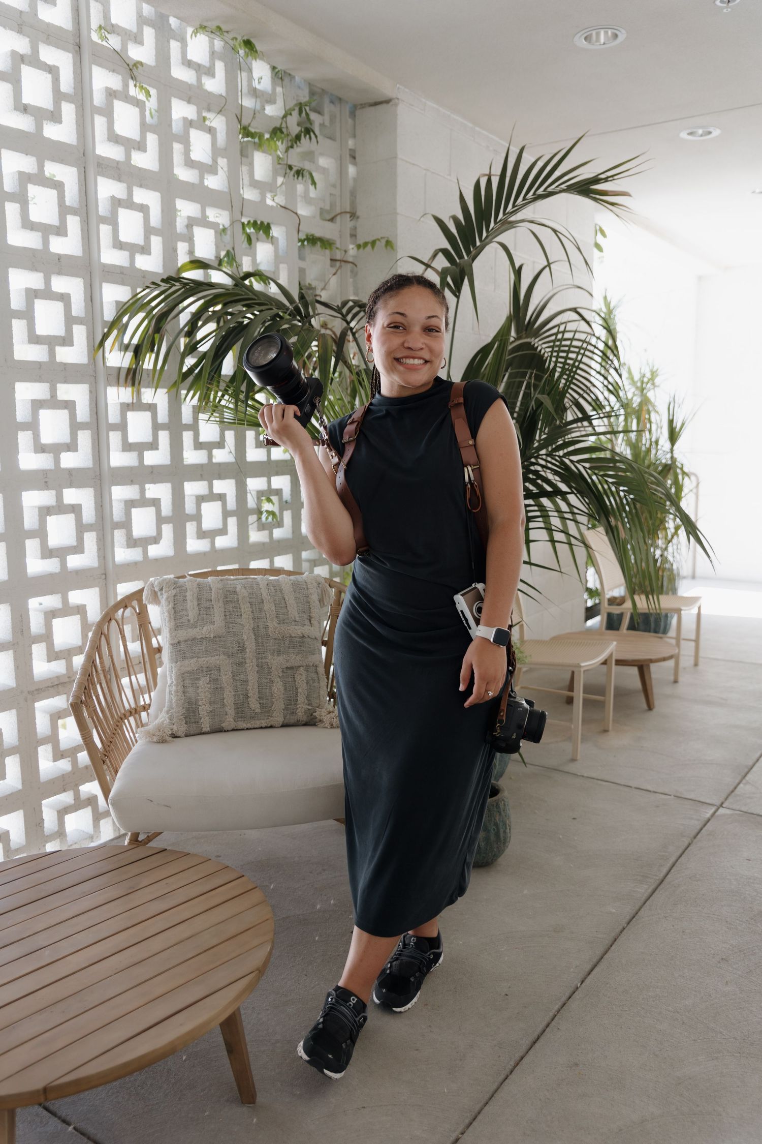 Person in black dress poses playfully in a modern white hallway with geometric wall details and potted plants.