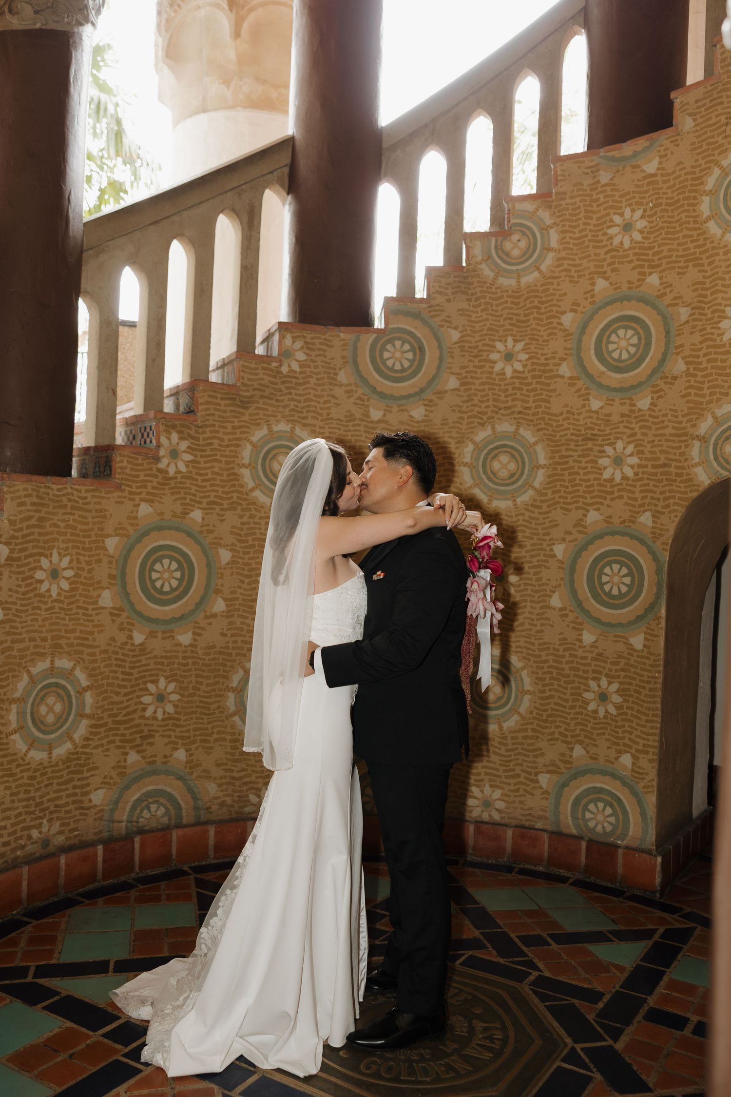 Bride and groom share first kiss in ornate tiled rotunda with arched windows during elegant wedding ceremony.