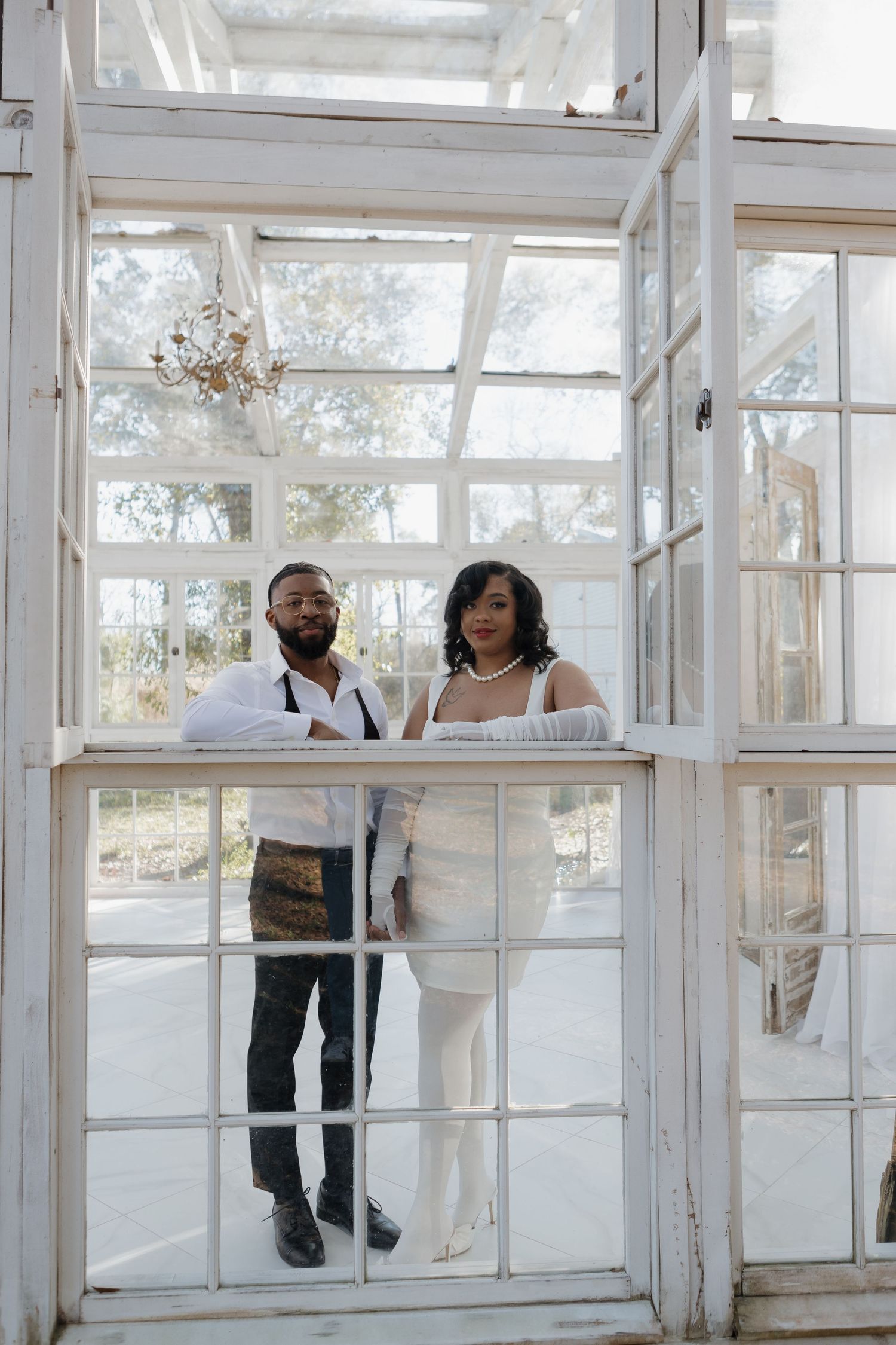 Couple poses together in a bright sunlit greenhouse with white window frames.