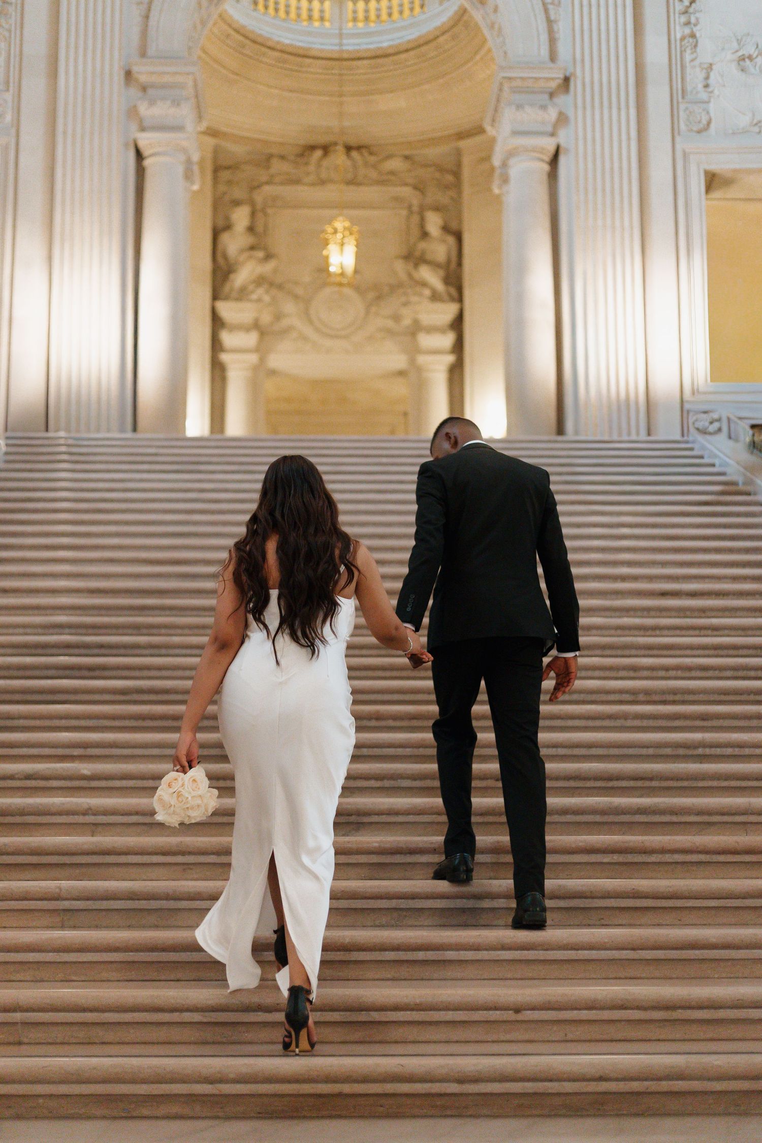 A couple ascends grand marble stairs in formal attire.