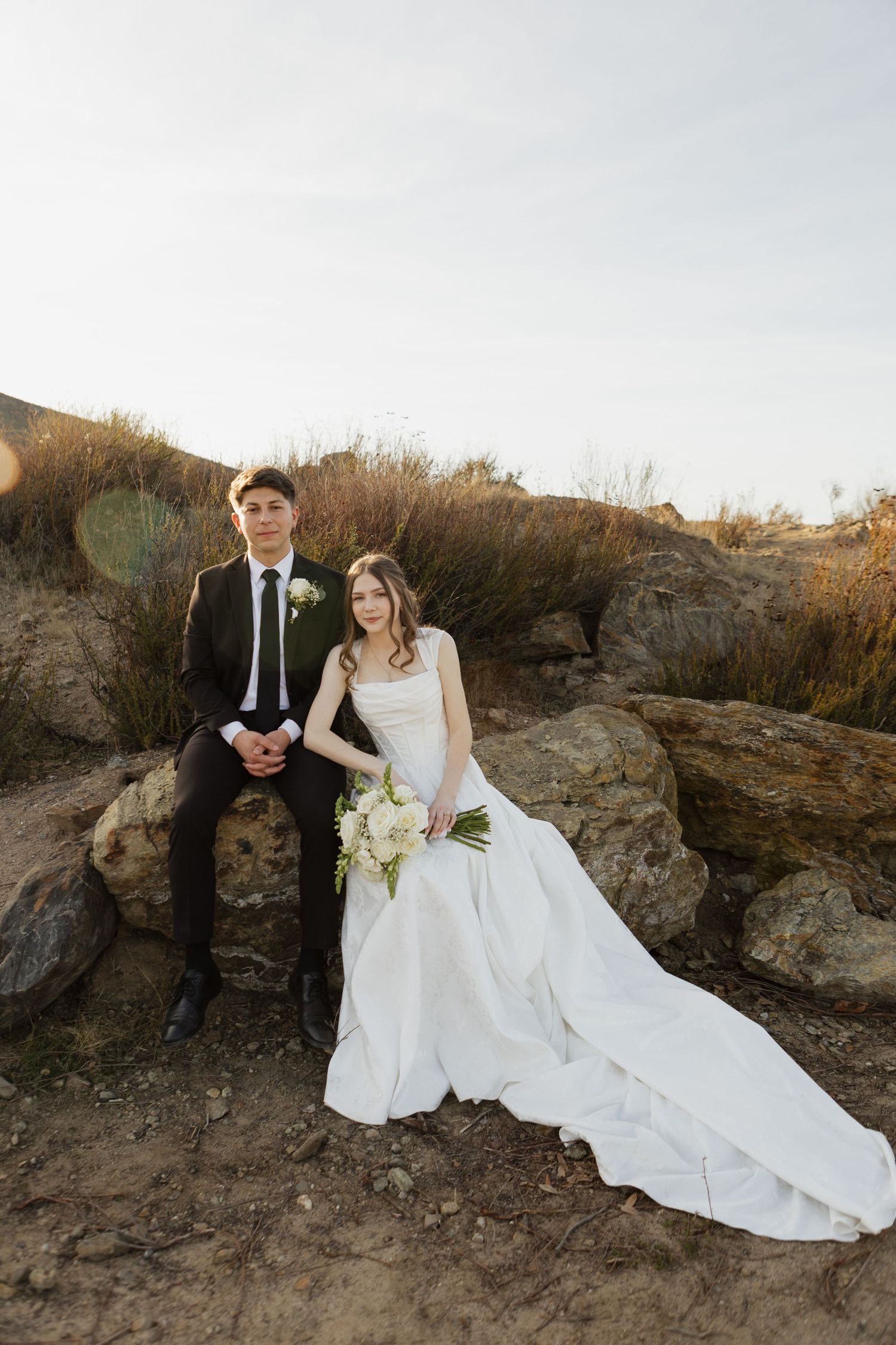 A wedding couple sits on rocky terrain with the bride's flowing white dress trailing behind.