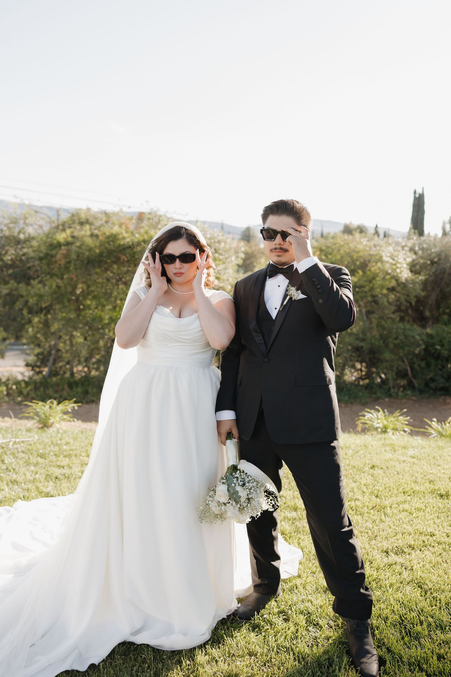 Bride in white gown and groom in black tuxedo posing outdoors in sunglasses, holding bouquet on green lawn.