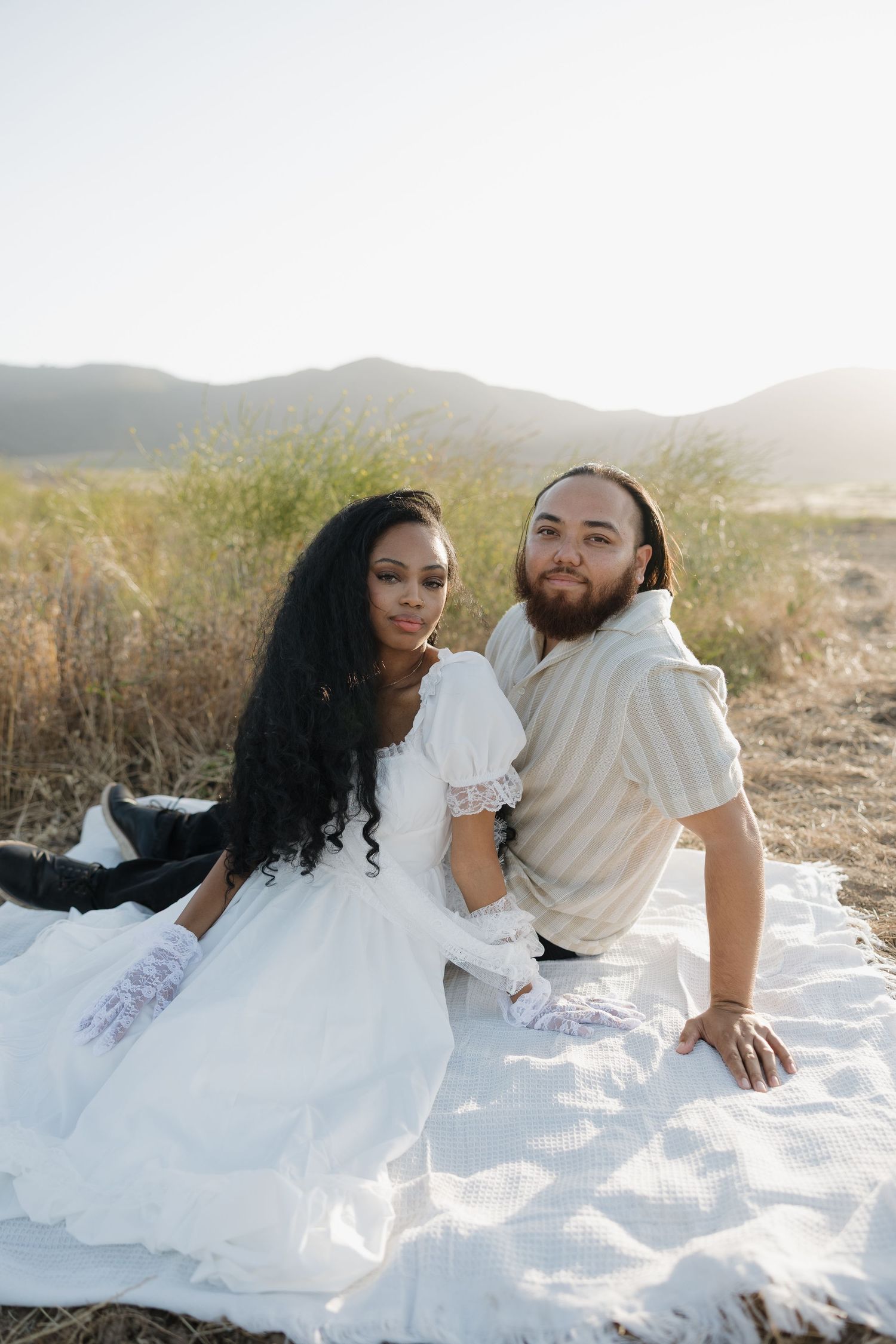 couple sits on a white blanket in a sunlit field with mountains, woman in white dress, man in striped shirt. Southern California photography