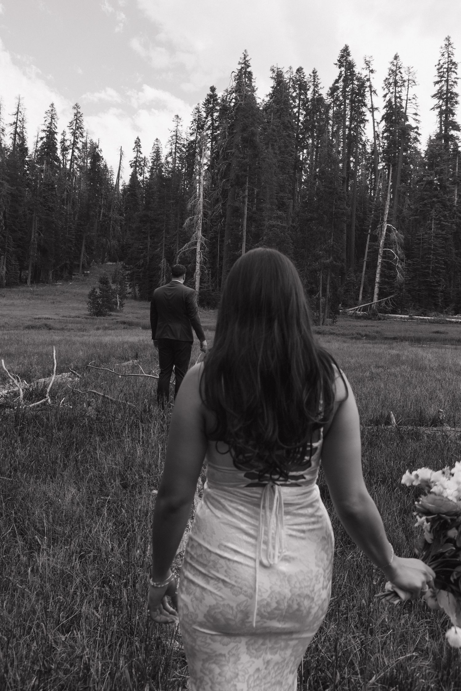 A couple walks through a forest meadow with tall pine trees in the background.