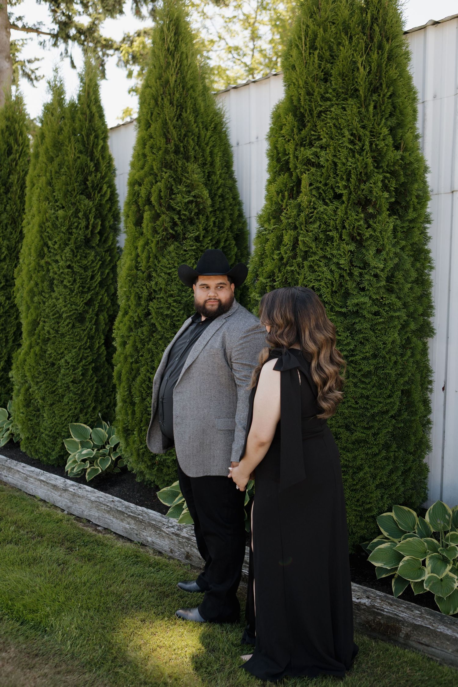 Two people in dark formal wear stand near a row of evergreen trees against a white fence.