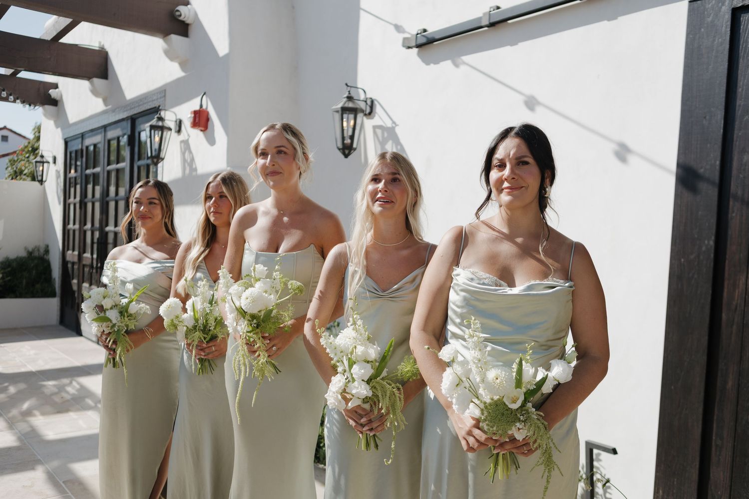 Bridesmaids in cream dresses hold bouquets while standing in bright sunlit space.