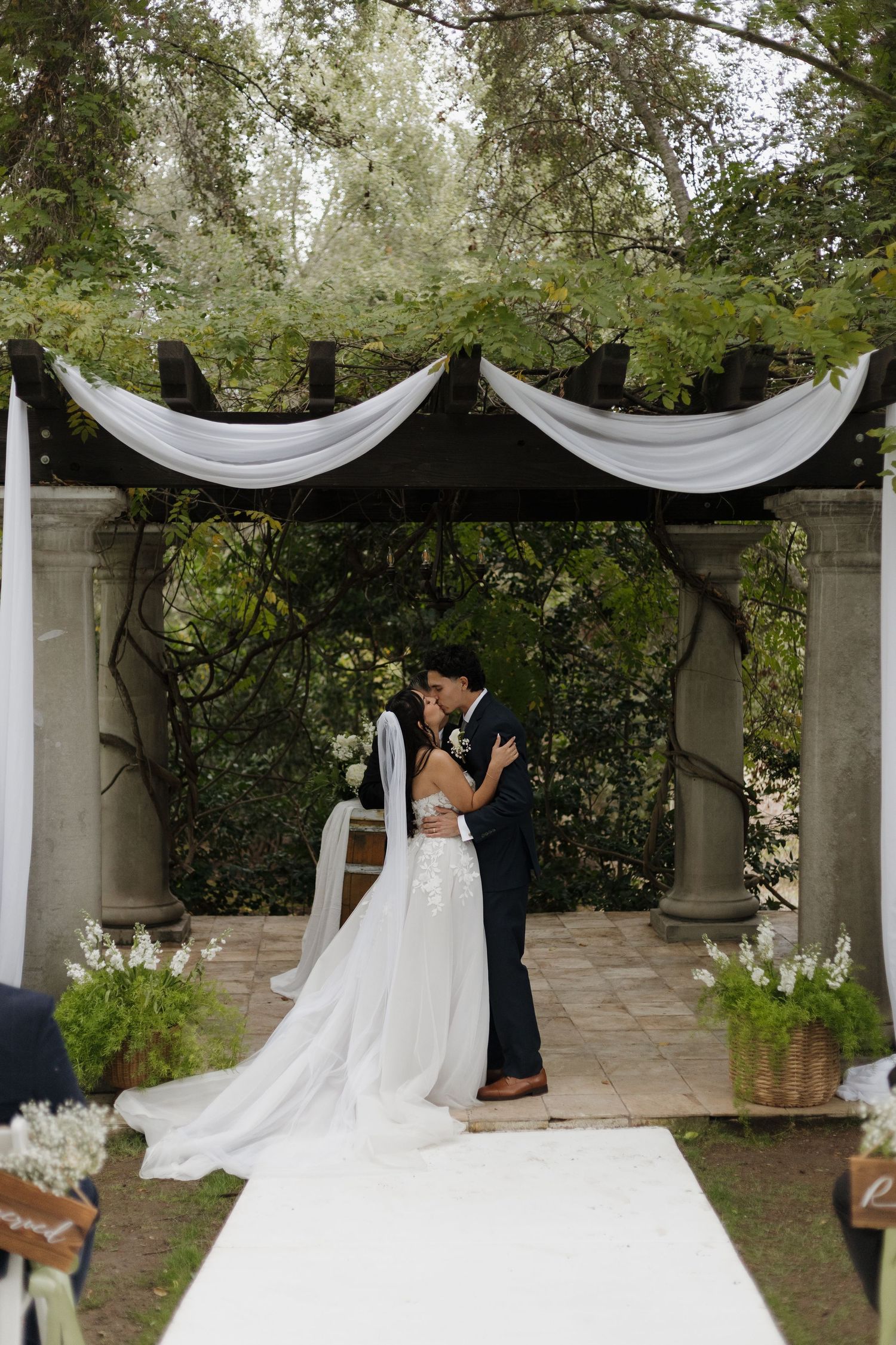 A romantic wedding ceremony under a white draped canopy surrounded by lush greenery and stone columns.