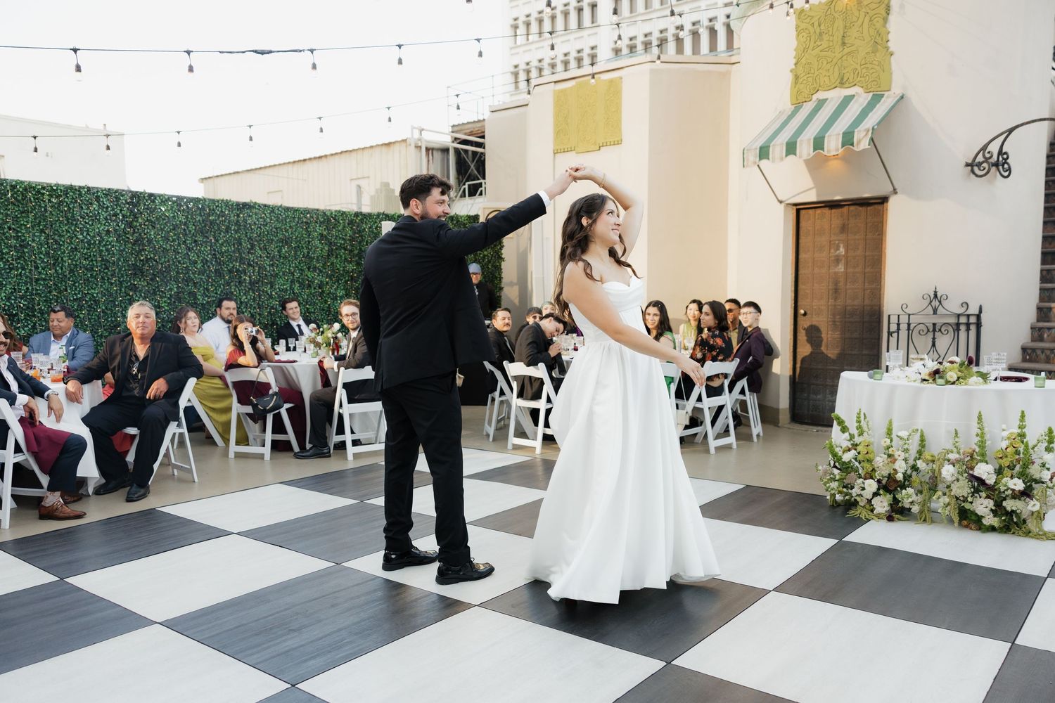 Bride and groom share first dance on checkered floor at outdoor wedding reception with seated guests.