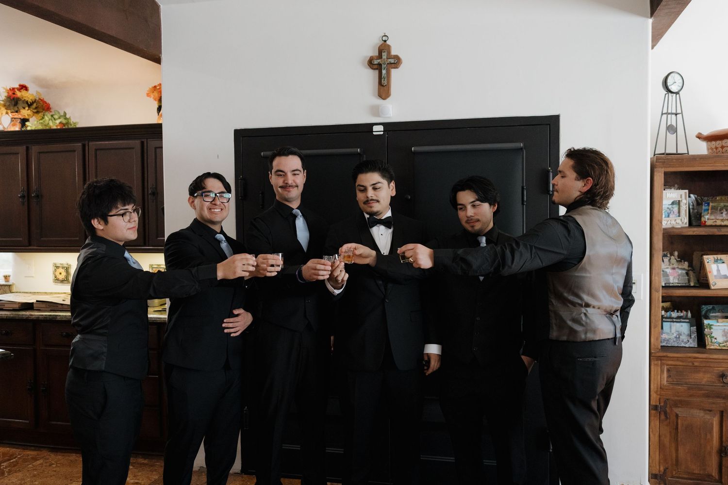 Groom and groomsmen in black tuxedos laughing together indoors before wedding ceremony.
