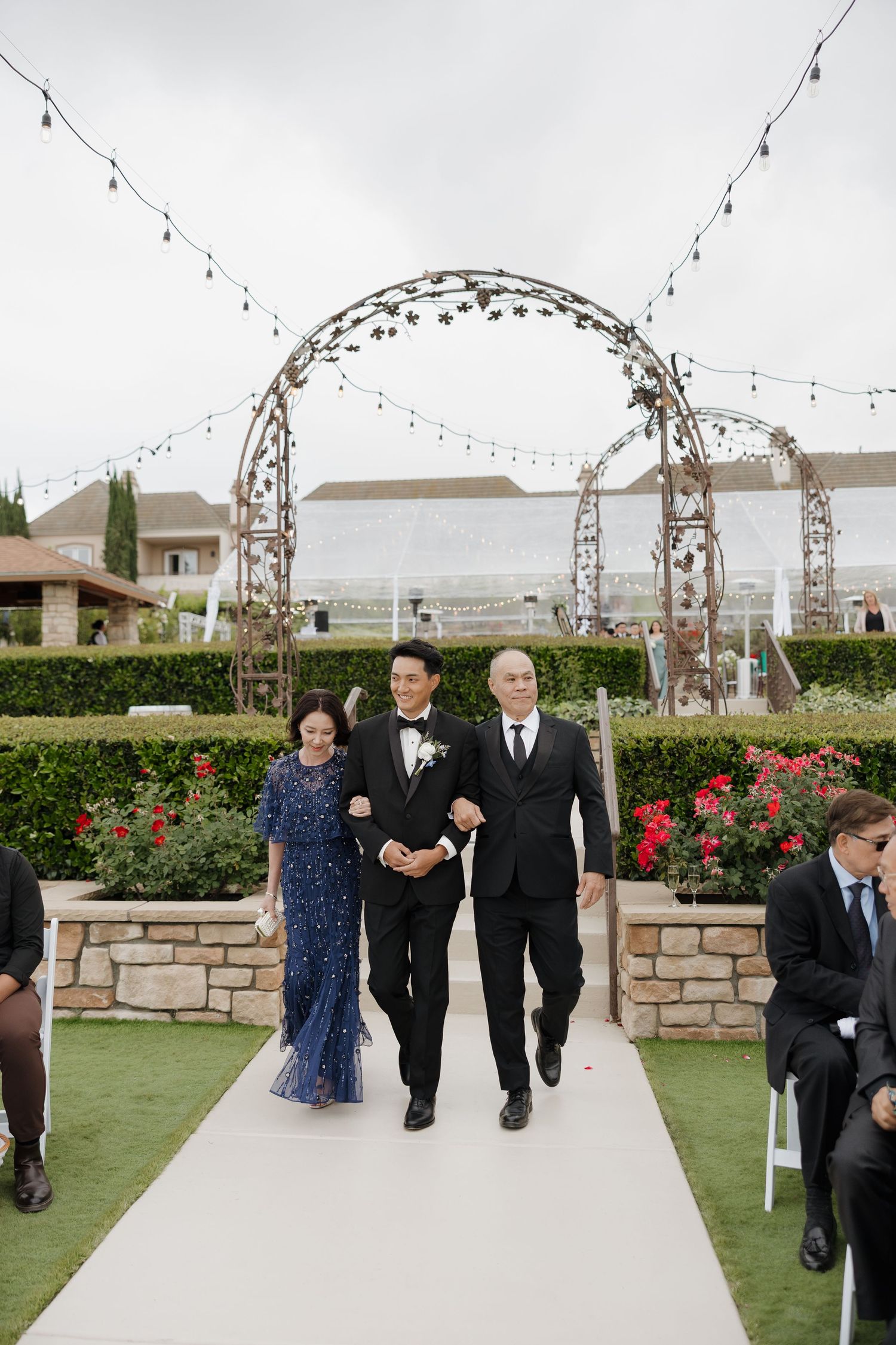 Groom in black tuxedo walks down outdoor wedding aisle escorted by parents, with floral arch and string lights backdrop.