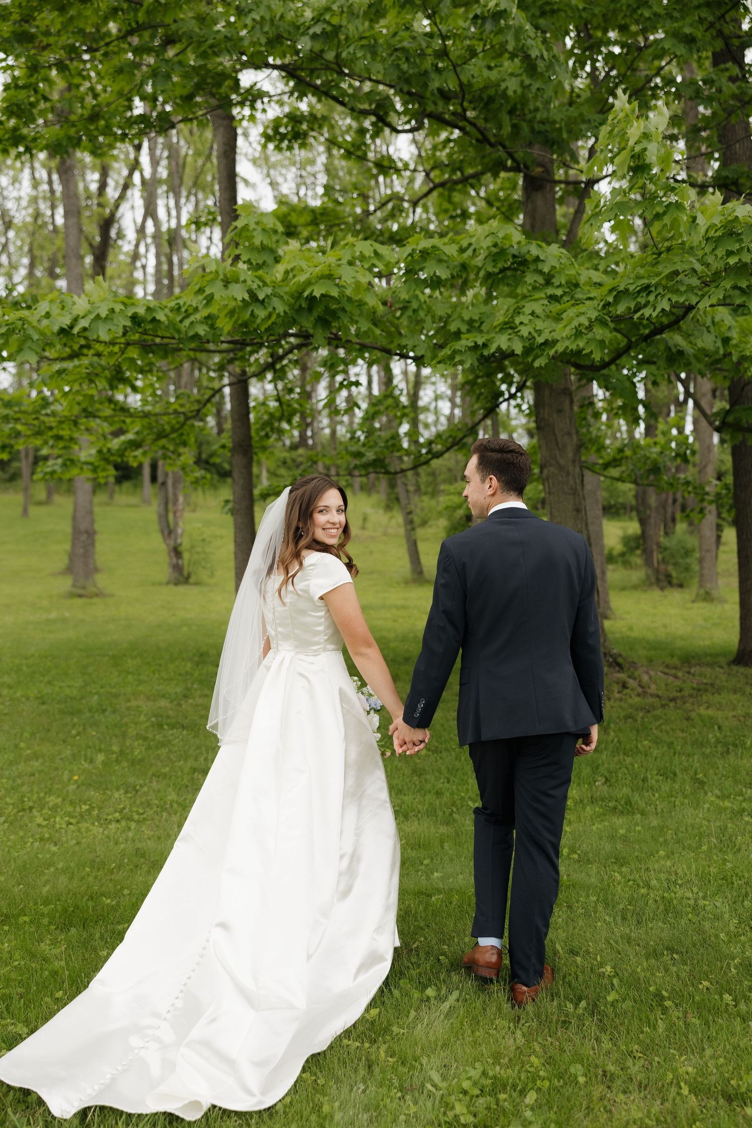 Bride and groom walking hand in hand through lush green park, bride in white gown with veil looking back smiling.