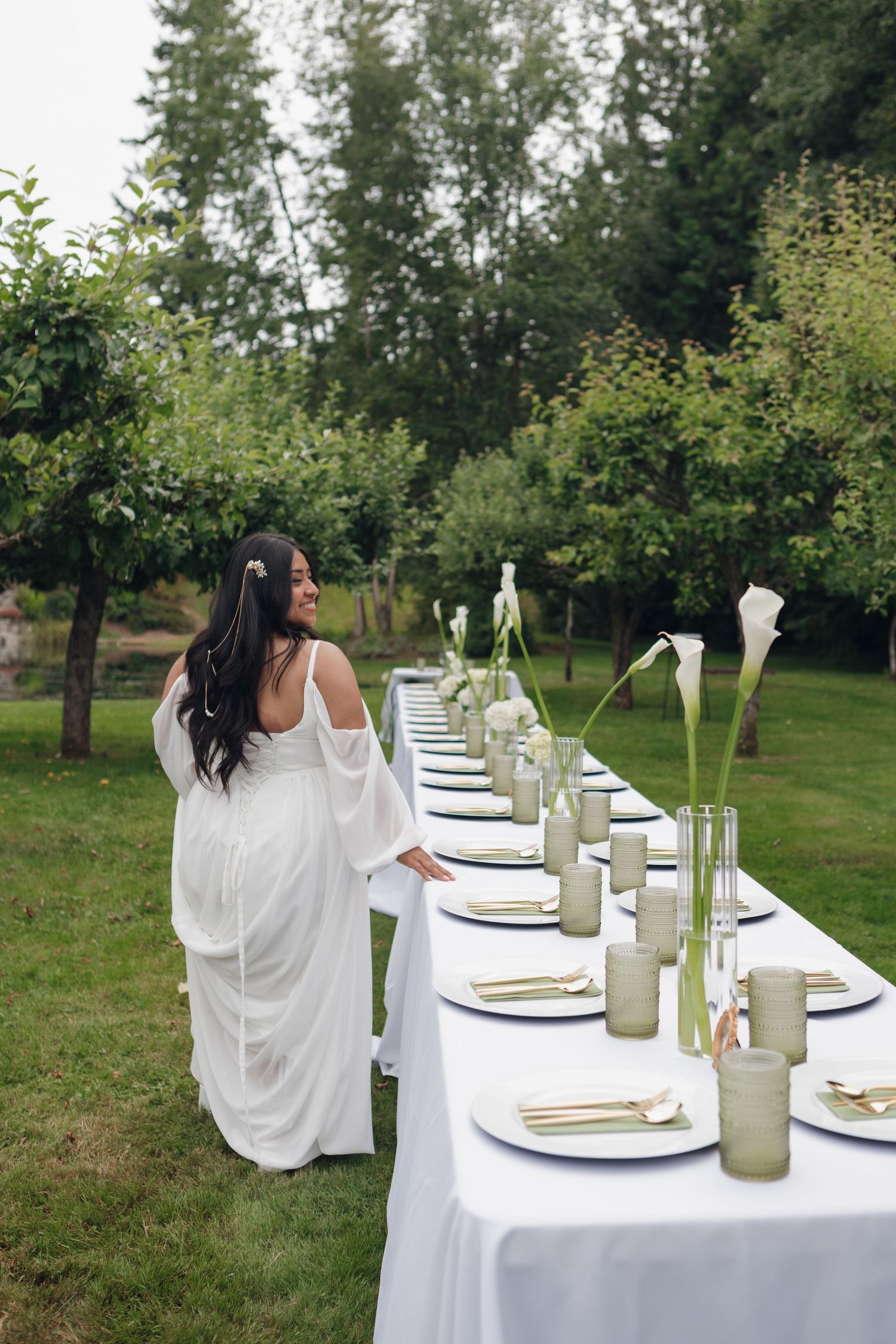 Elegant white outdoor table setting with place settings and glass bottles in a garden.