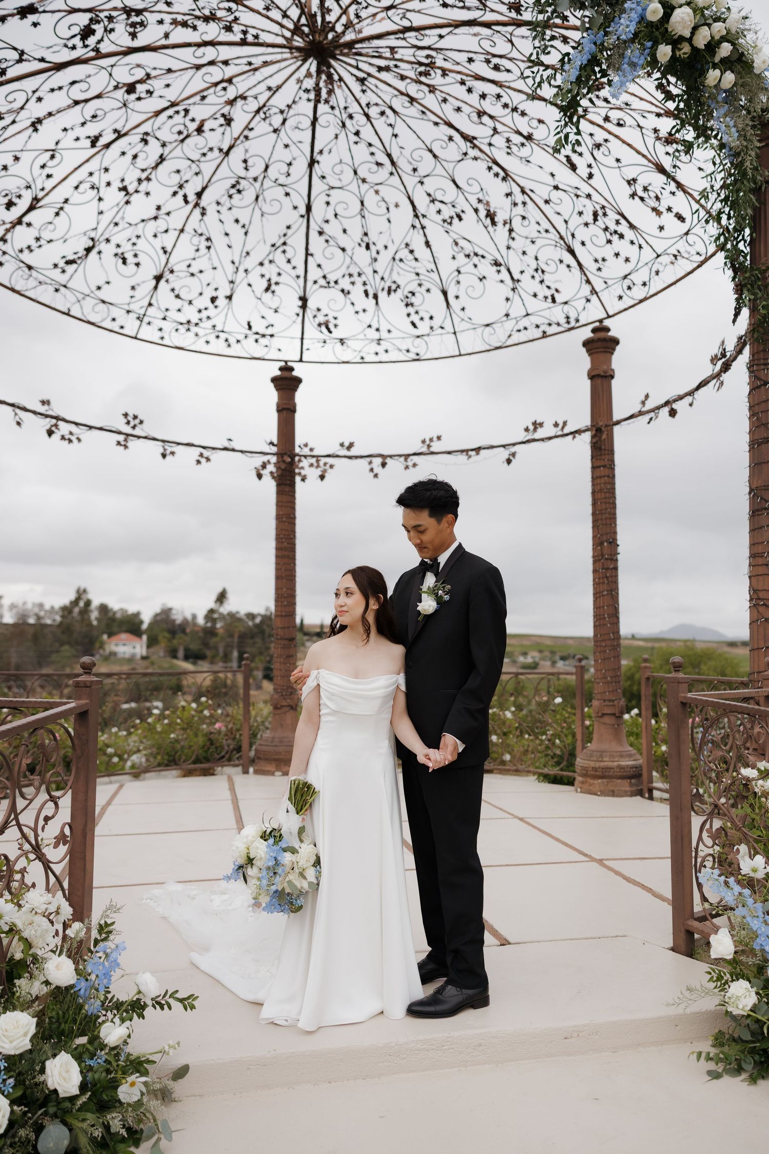 Bride and groom pose under ornate iron gazebo, she holds blue and white bouquet, overcast sky backdrop.