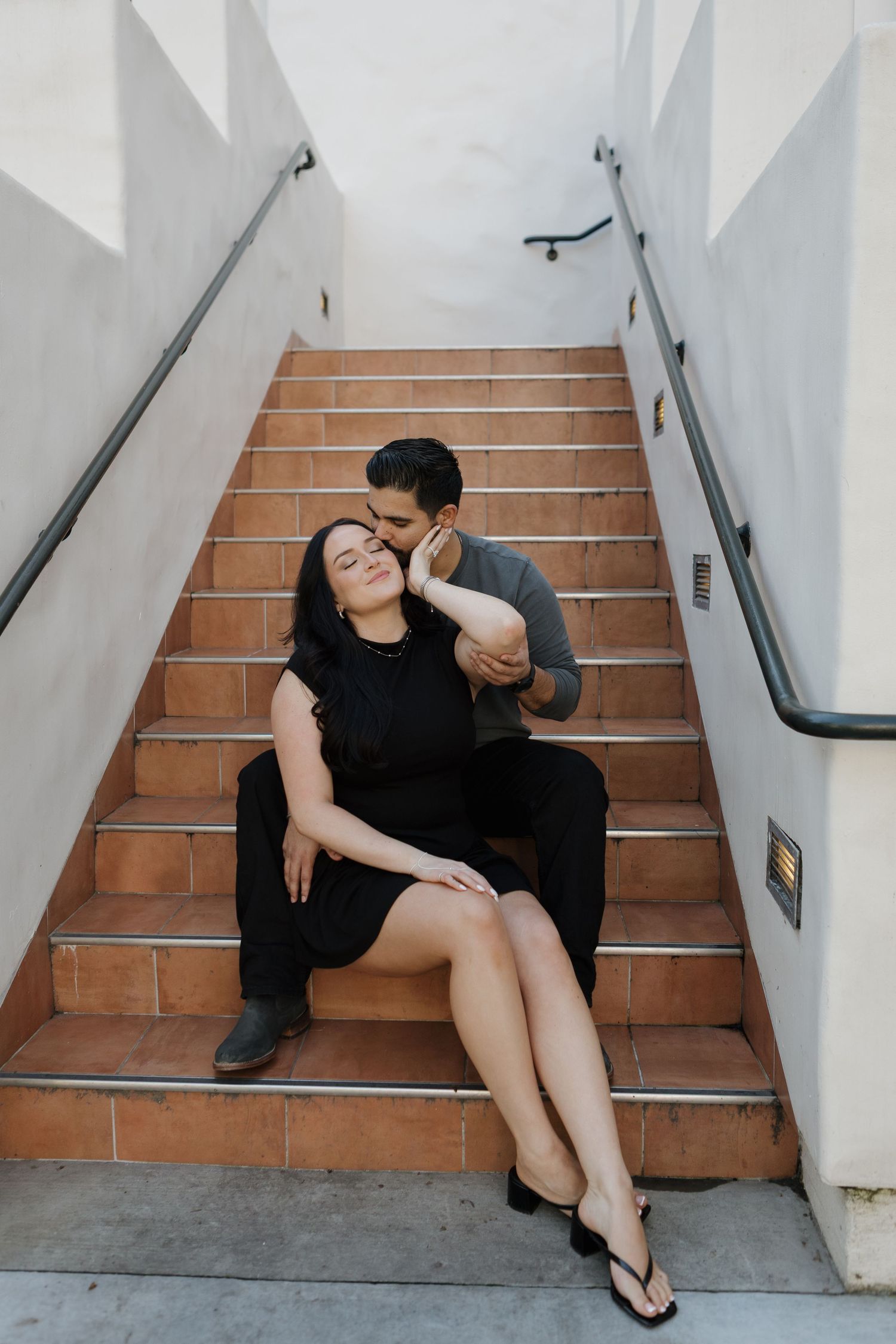 Couple in black outfits poses casually on terracotta stairs against white walls.