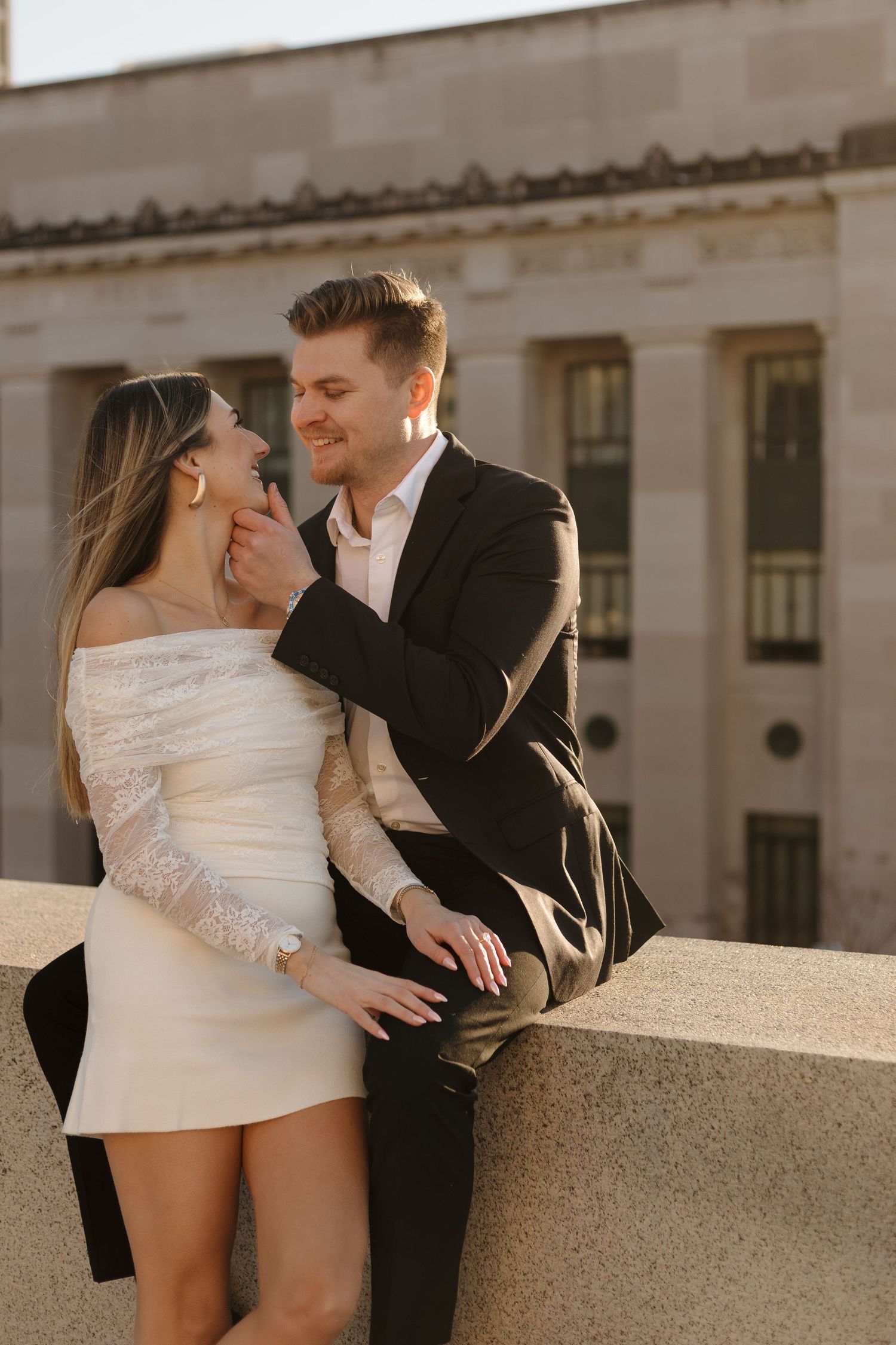 Romantic couple embracing against stone building wall in formal attire.