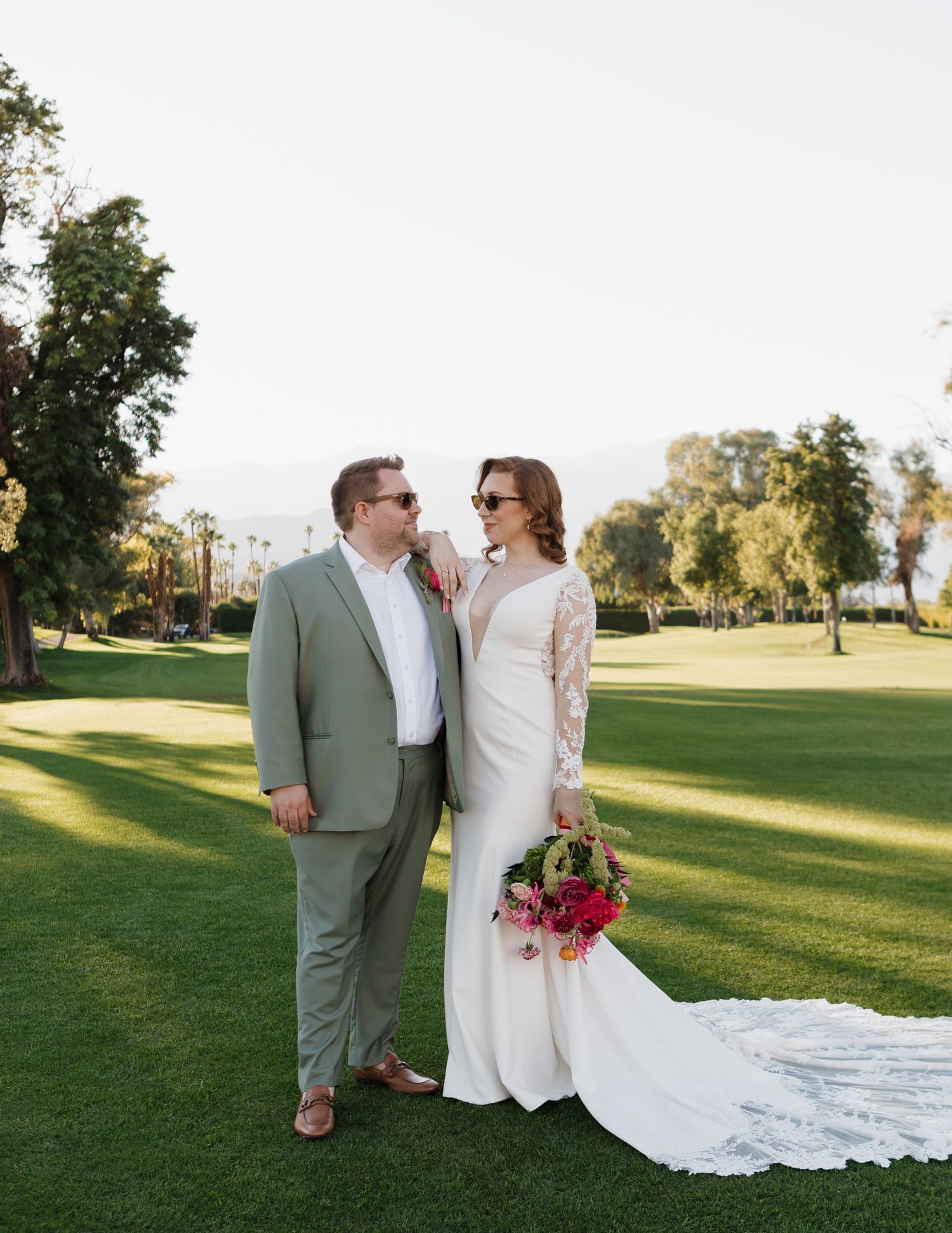Newlyweds pose together on a sunlit golf course with mountains in the background.